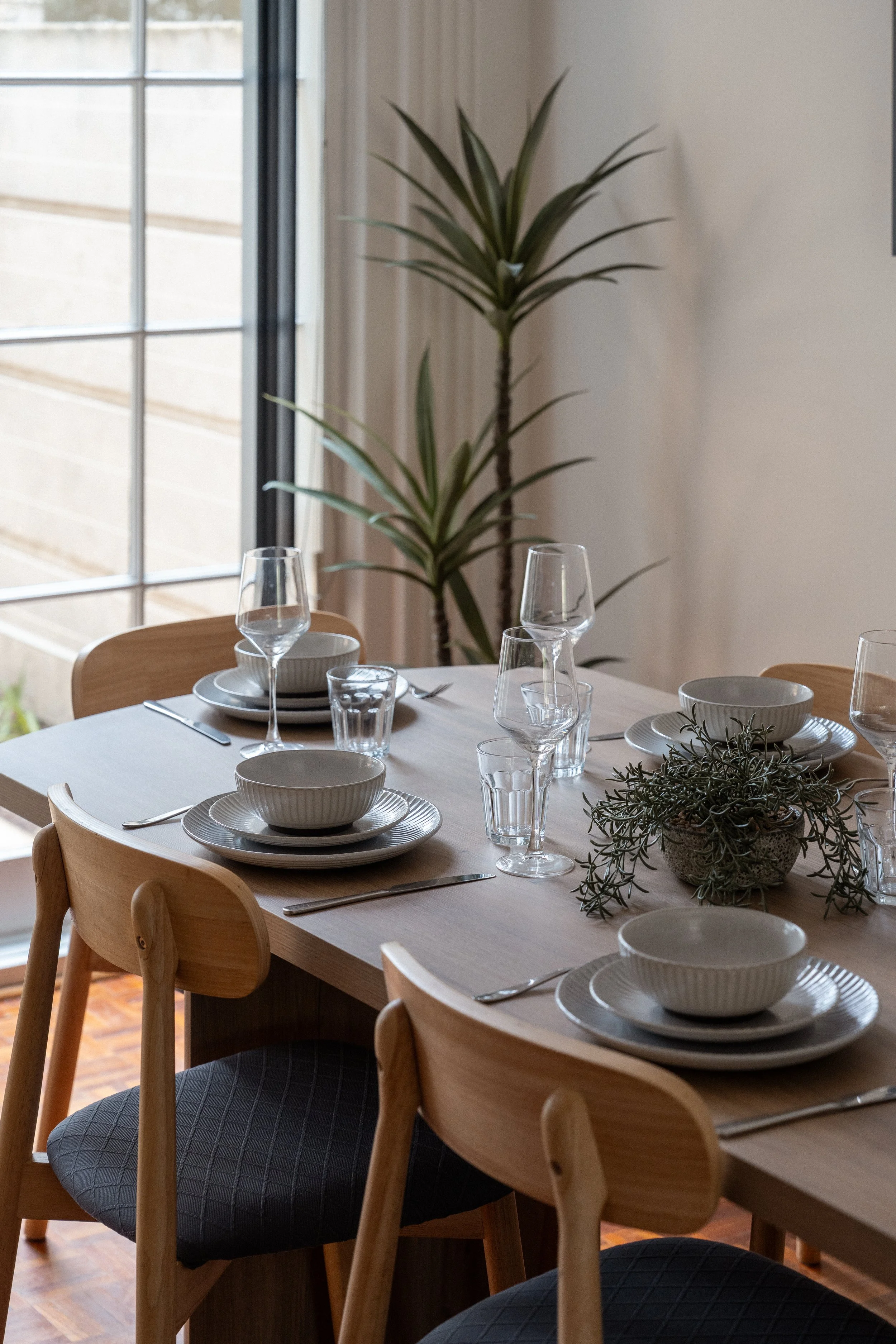 A dining table set with white bowls, glassware, and silverware, surrounded by wooden chairs with black cushioned seats, near a window in a well-lit room with a potted plant in the background.