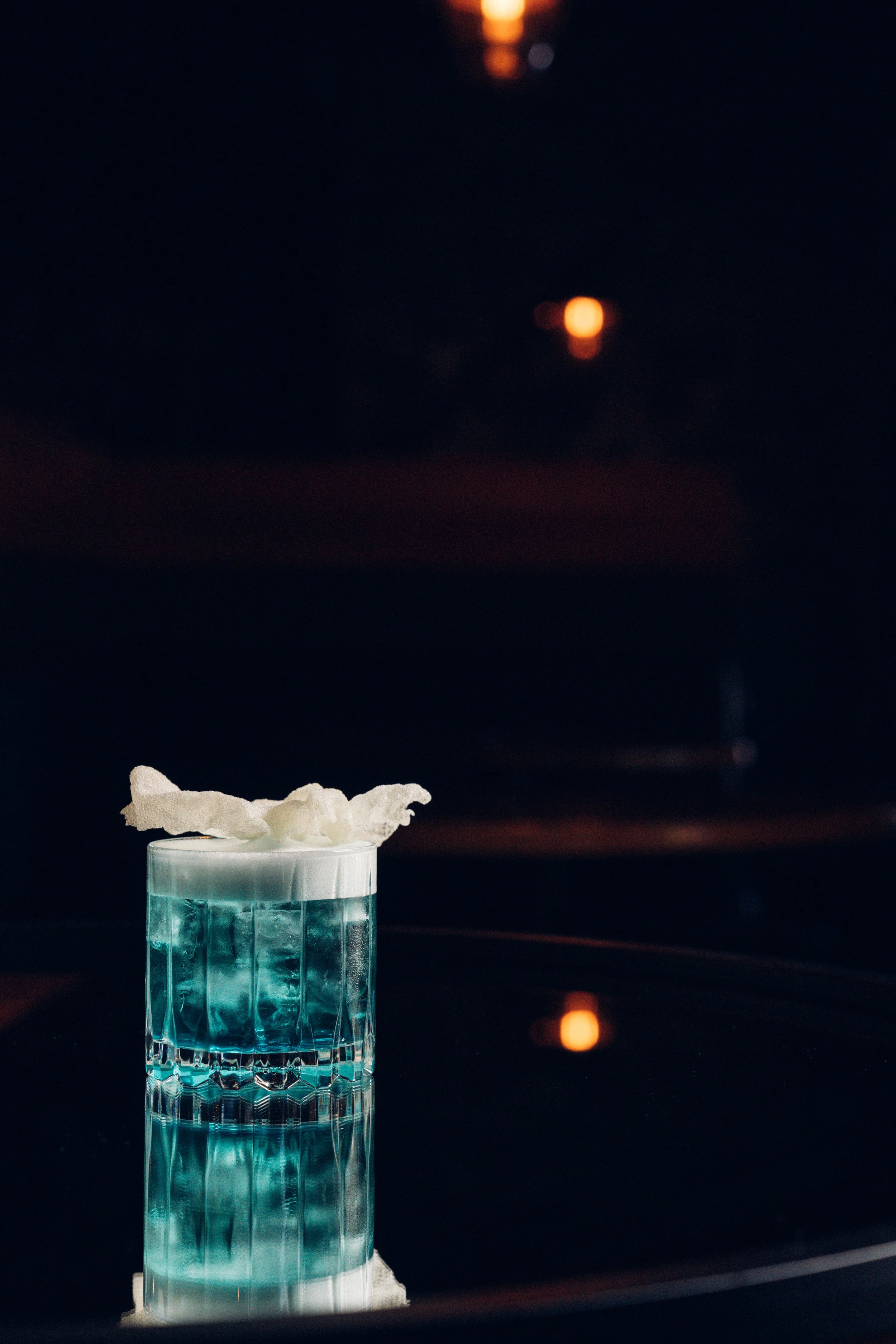 A glass with a bright blue drink topped with whipped cream, placed on a reflective surface with a scenic sunset view in the background.