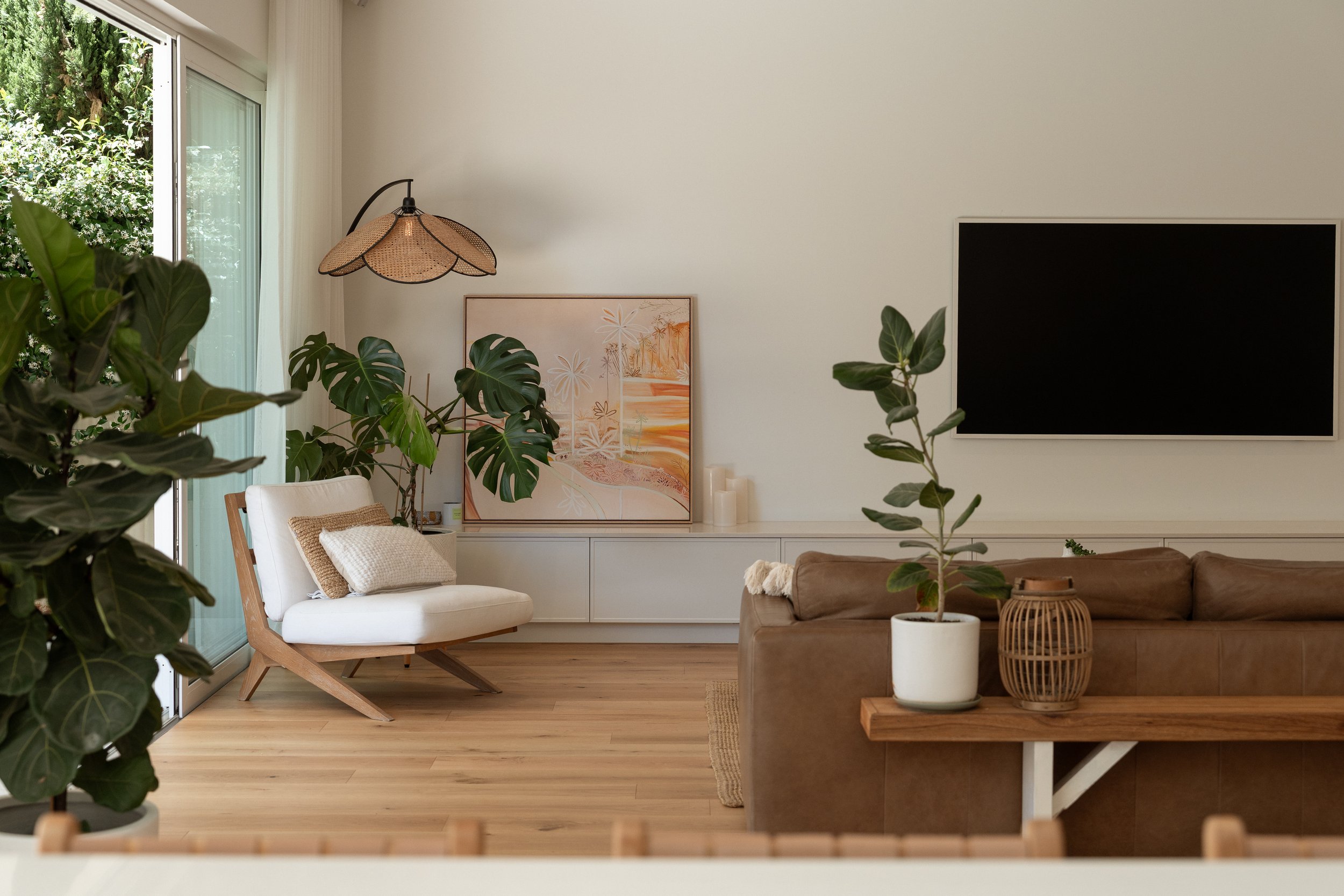 Bright living room with large sliding glass door, wooden flooring, and green houseplants. Includes a white armchair with beige cushions, a brown sofa, a flat-screen TV on the wall, and decorative art and candles.