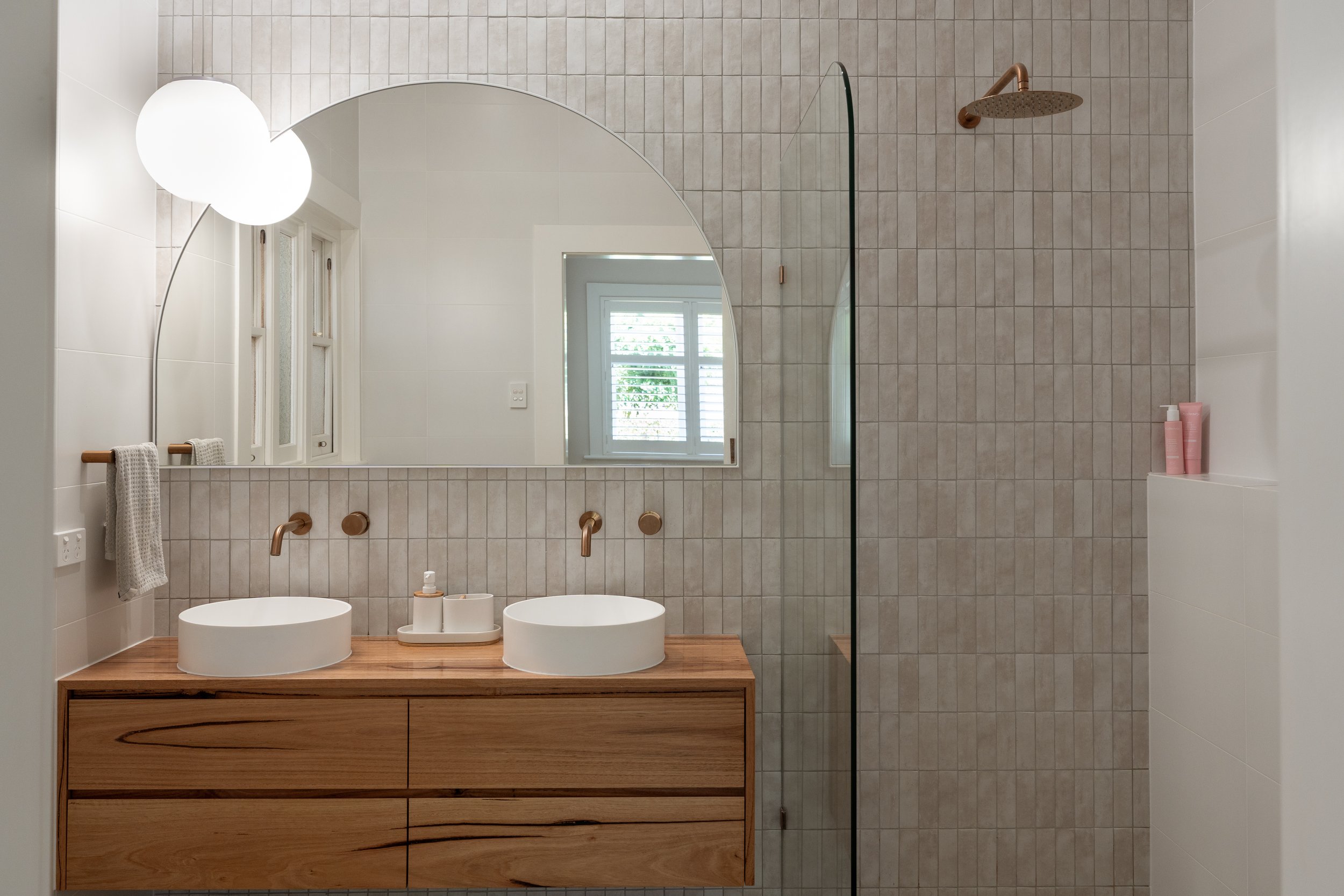 Modern bathroom featuring a wooden vanity with double vessel sinks, round mirror, copper fixtures, a large shower with a glass divider, a window with louvered blinds, and minimal decor.