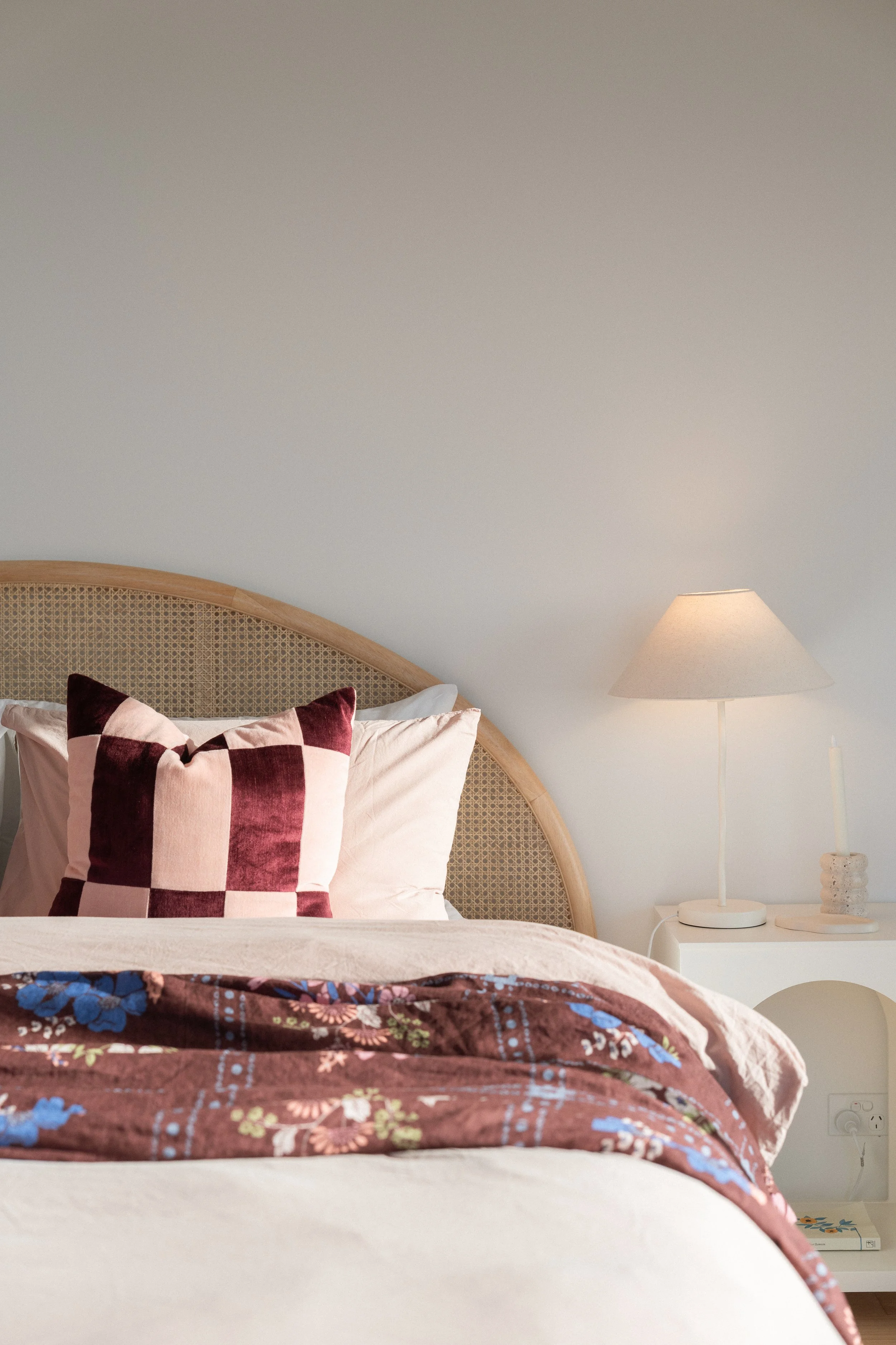 A bedroom showing part of a bed with a quilted bedspread and a decorative pillow with a checkered pattern. A nightstand with a modern lamp and a small decorative object is beside the bed. The wall is painted in light, neutral color.