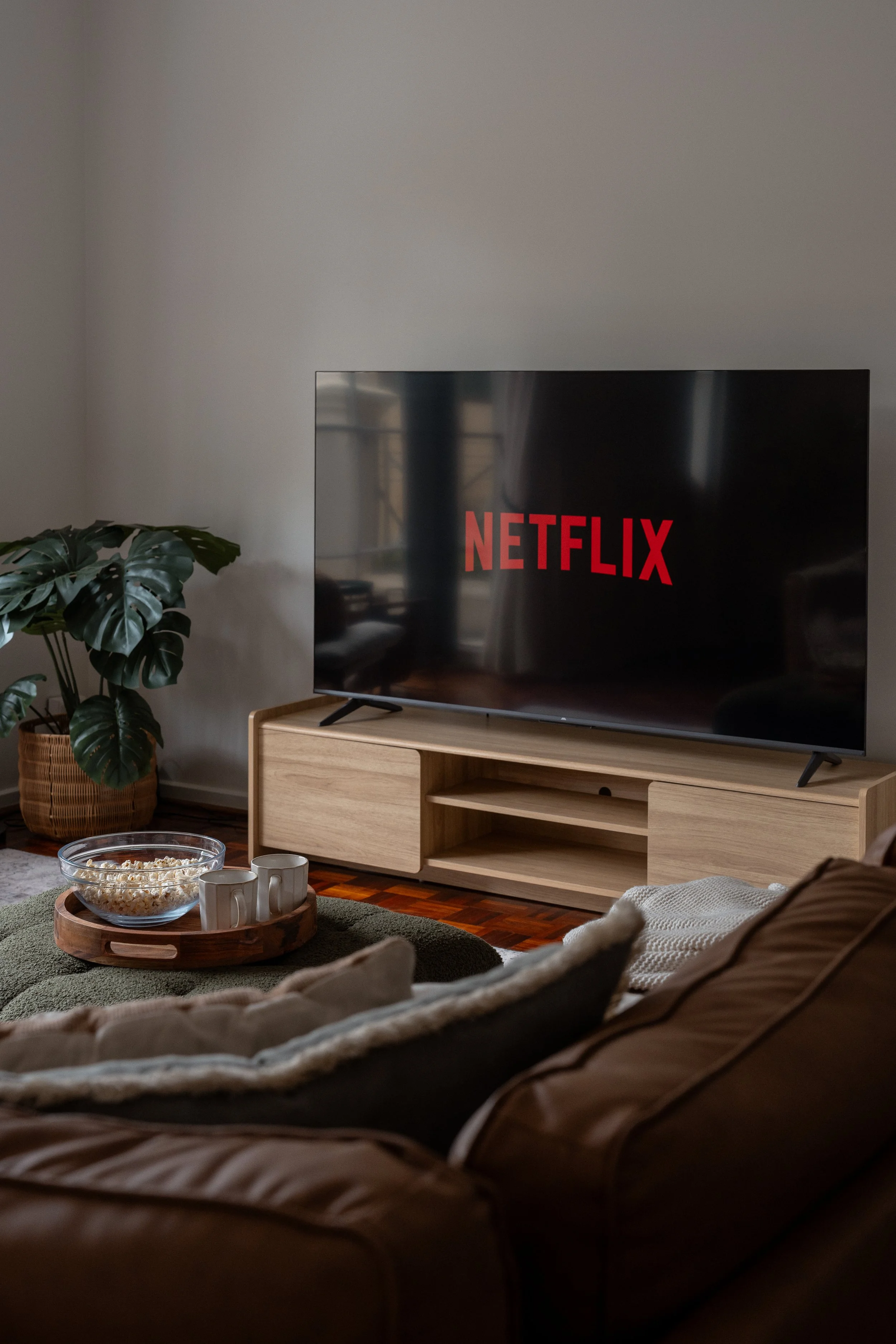 Living room with a flat-screen TV displaying the Netflix logo, a wooden TV stand, a potted plant, a bowl of popcorn and two coffee mugs on a tray, and a couch with a blanket in the foreground.