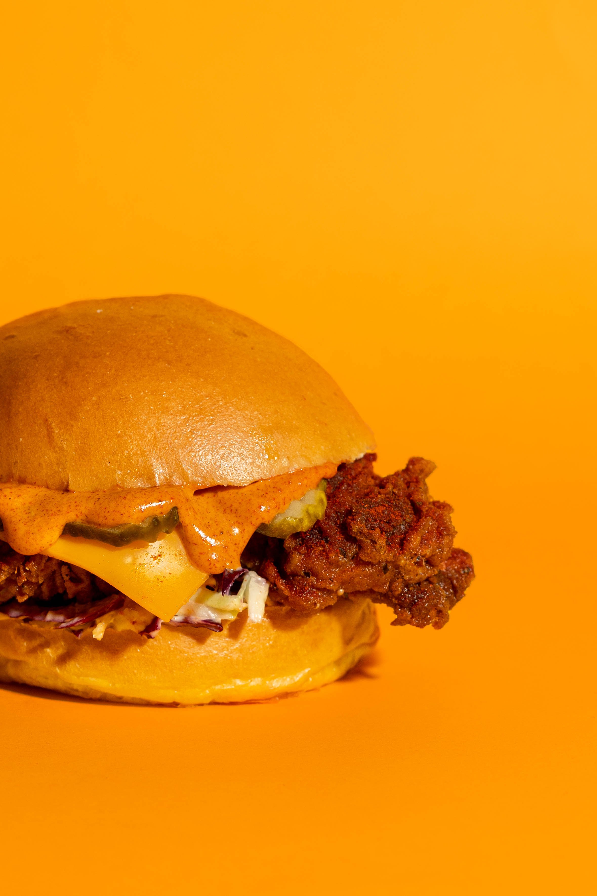 Close-up of a cheeseburger with fried chicken patty, cheddar cheese, lettuce, pickles, spicy sauce, and a bun against an orange background.