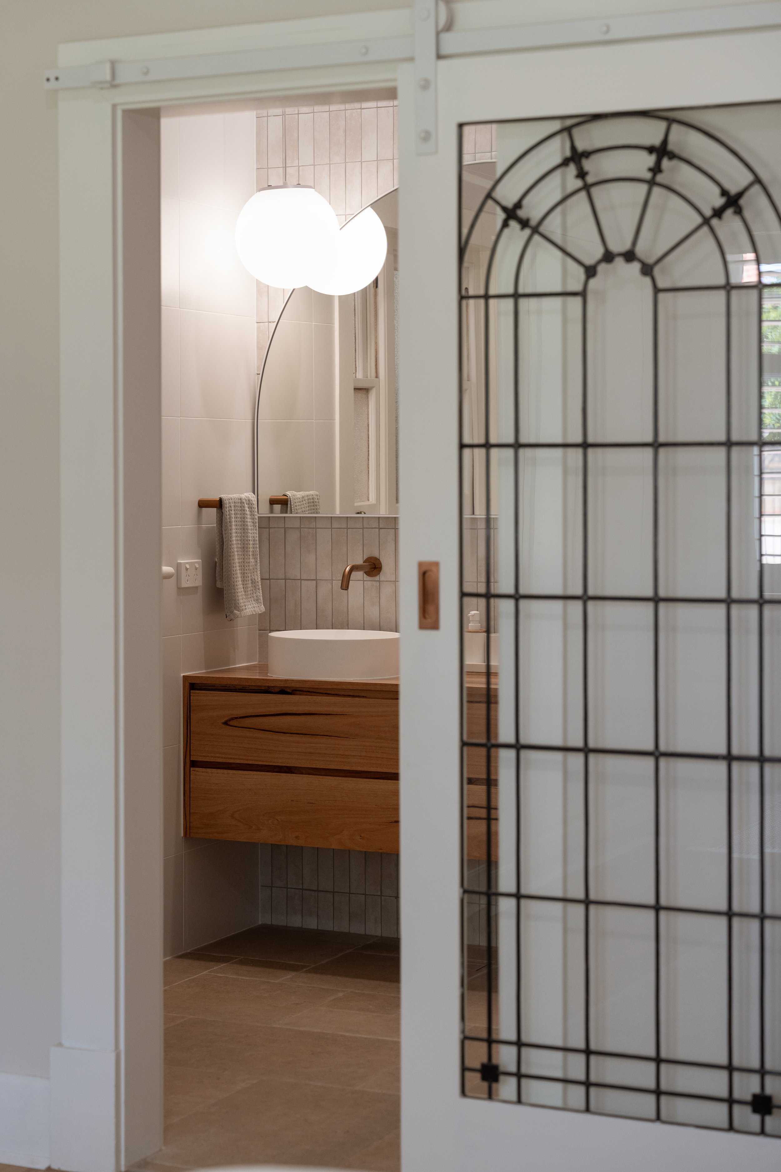 View through a sliding door into a modern bathroom featuring a round vessel sink, wooden vanity, beige tiles, and a tall curved floor lamp with white bulbs.