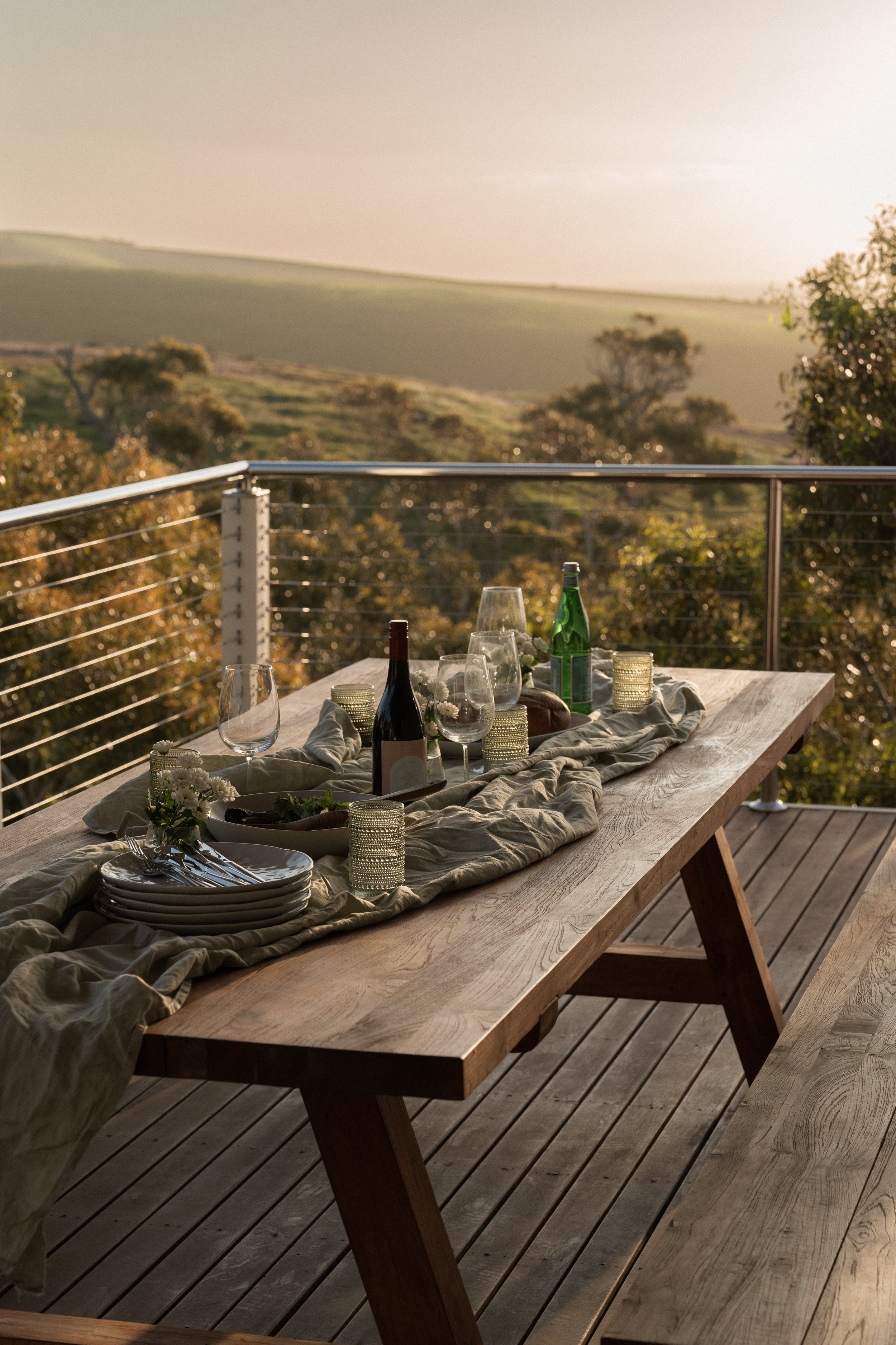 Outdoor dining table set on a wooden deck with wine bottles, glasses, plates, and candle holders, overlooking a scenic hilly landscape during sunset.