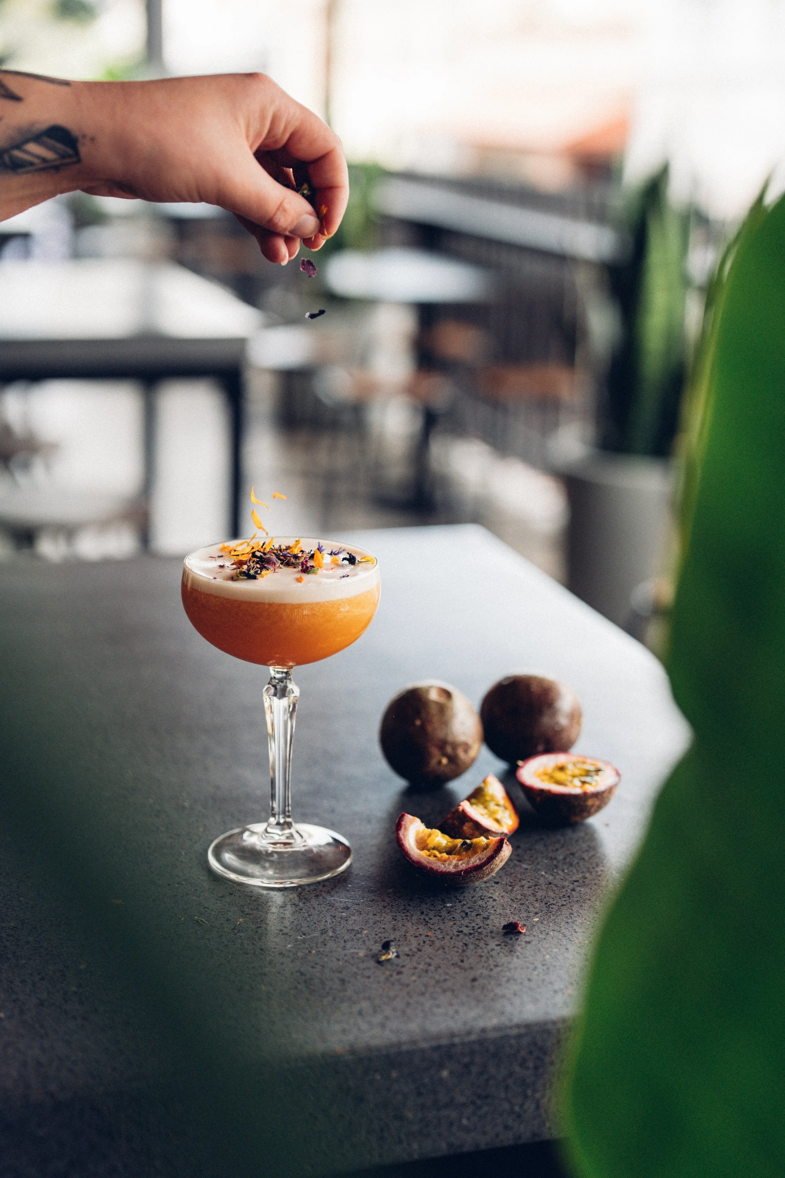 A hand is sprinkling edible flower petals over a Passion Fruit cocktail drink in a tall glass with a long stem. There are fresh passion fruits, some sliced open, on a black countertop near the glass. The background is blurred with greenery and indoor