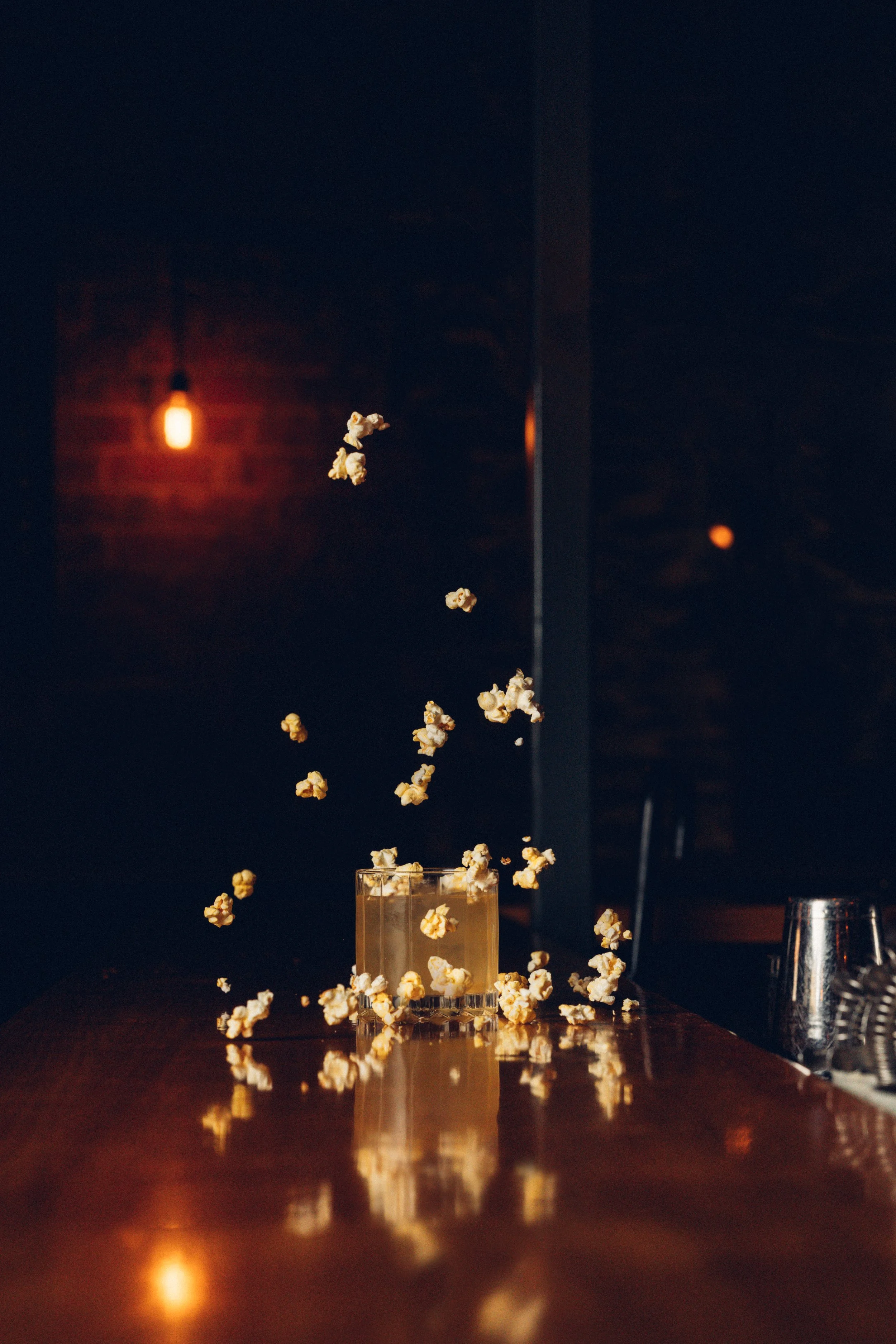 Popcorn falling into a glass on a wooden table at night, with warm lighting and a dark background.
