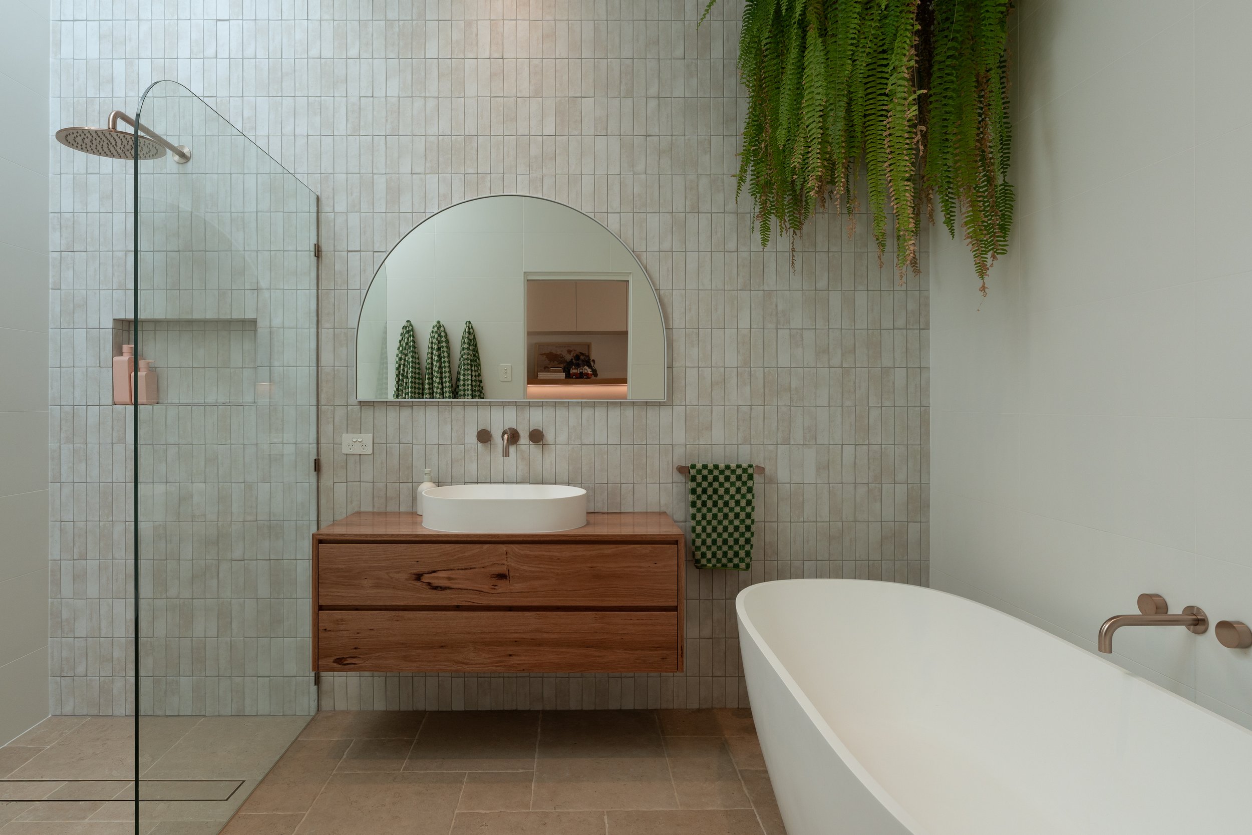 Modern bathroom with a walk-in shower, a wooden vanity with a round vessel sink, a large arched mirror, hanging green towels, a freestanding bathtub, and lush green plants hanging from the ceiling.