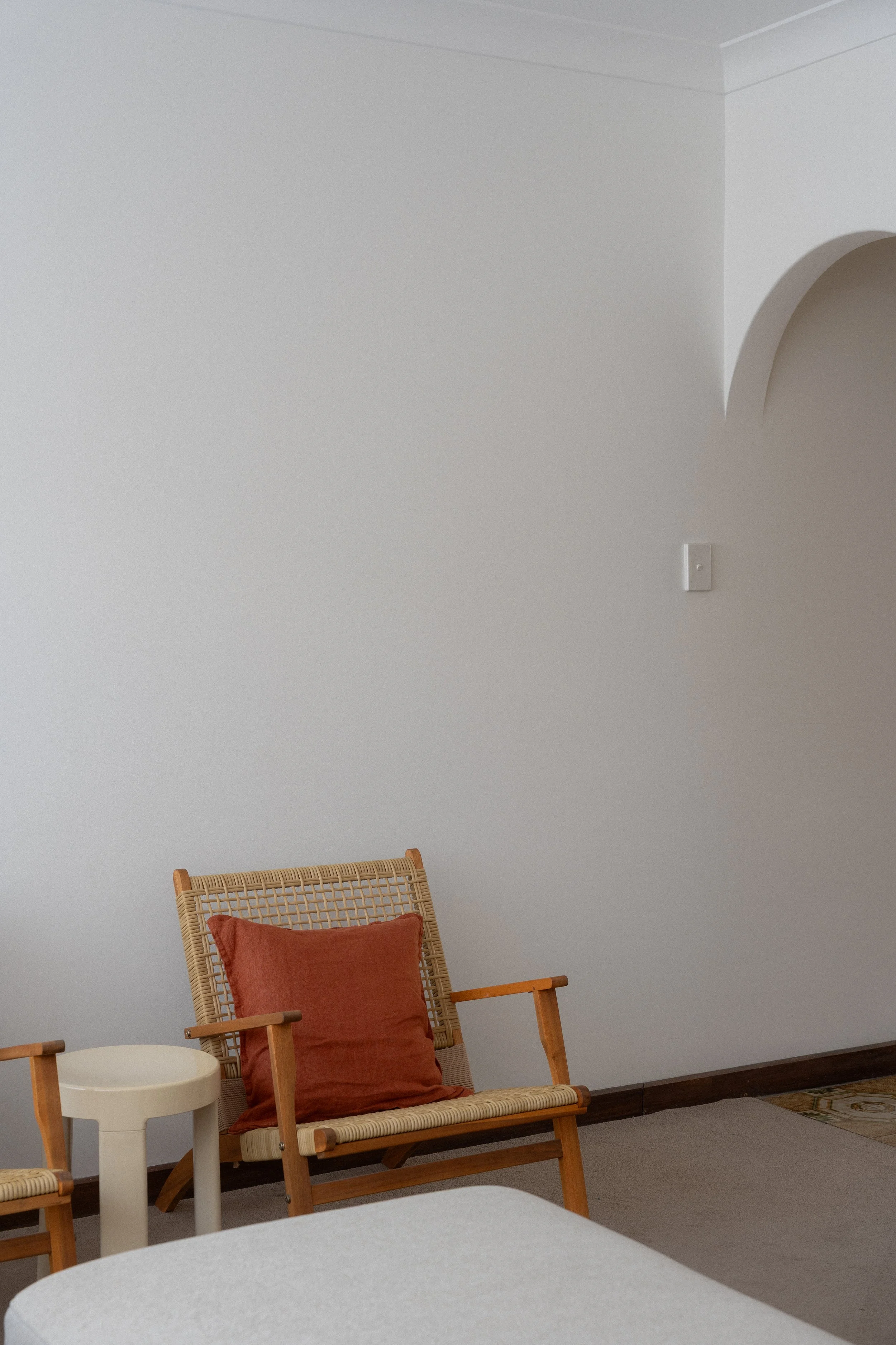 A beige rocking chair with a rust-colored cushion, next to a small white side table in a room with light gray walls, wood trim, and a patterned rug.