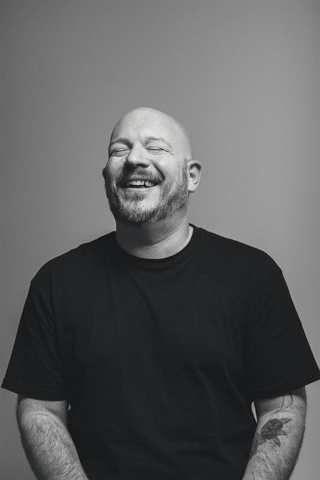 Black and white portrait of a smiling bald man with a beard, wearing a black T-shirt, standing against a plain background.