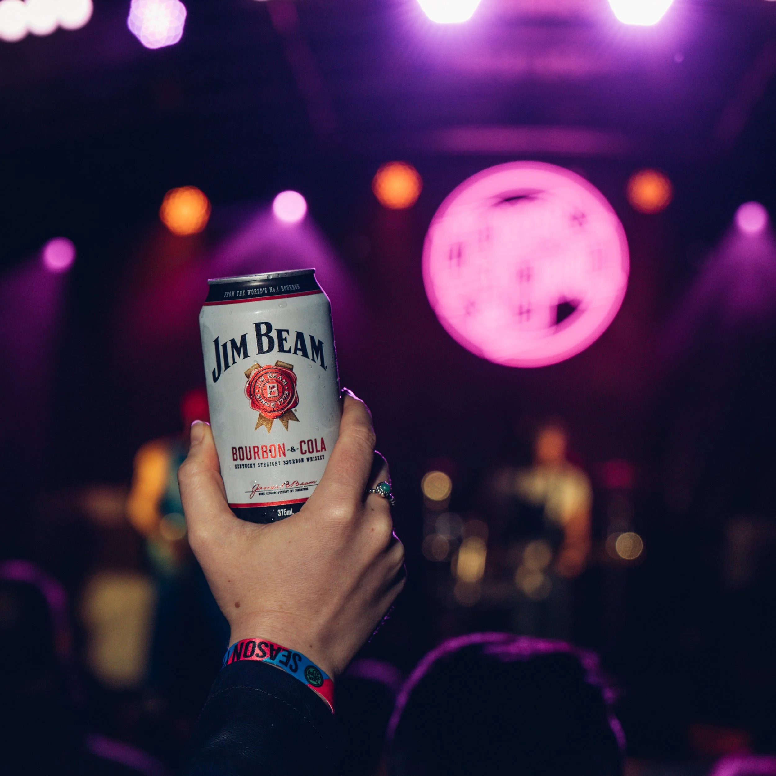 A hand holding a can of Jim Beam bourbon and cola against a blurred background of purple and pink stage lights, with a wristband that has 'SEASON' written on it.
