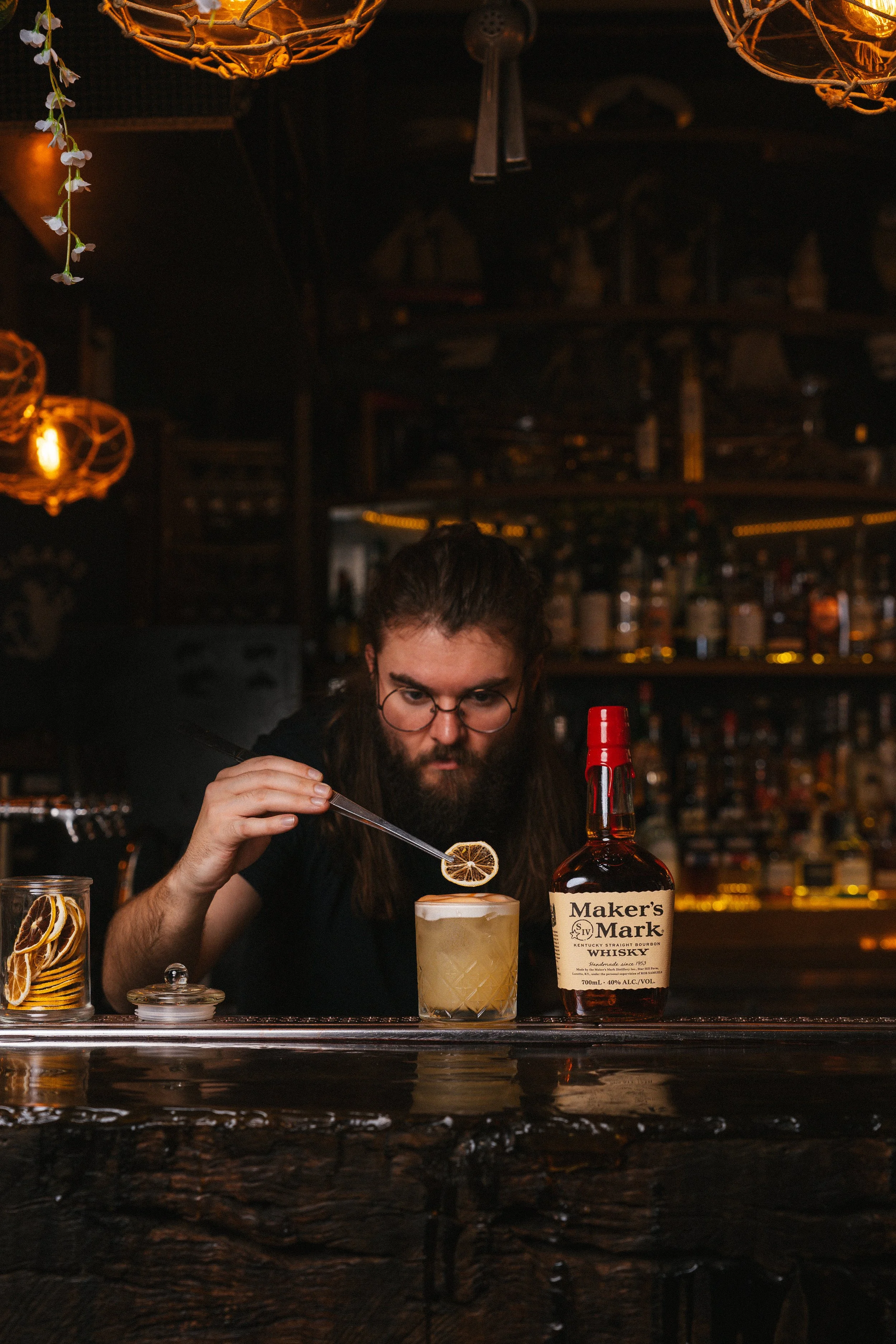 A man with long hair and glasses is preparing a cocktail at a bar, adding a lemon slice to the drink. There is a bottle of Maker's Mark whiskey on the bar, along with glass jars filled with lemon slices and dried lemon slices. The background features a warm, dimly lit bar with bottles and glasses.