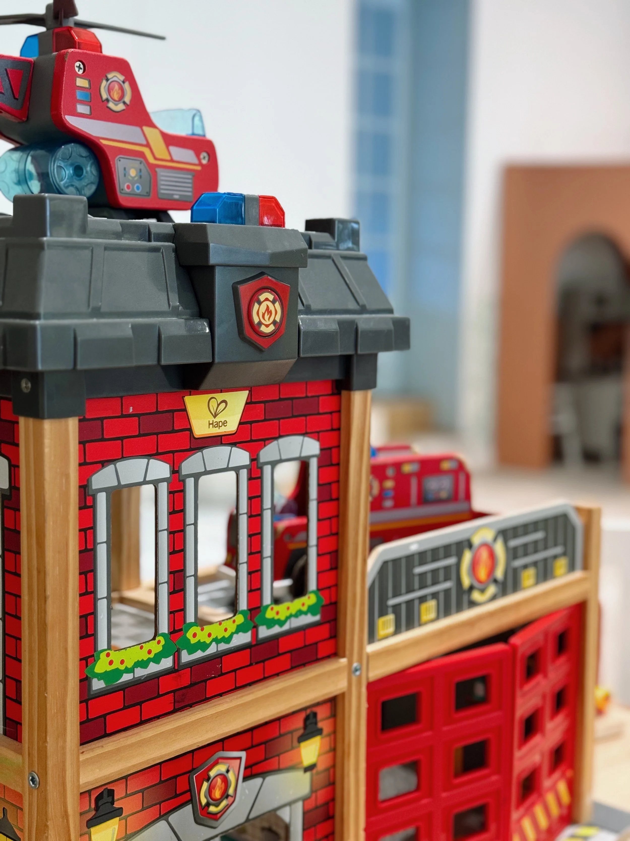 Une caserne de pompiers en jouet avec une voiture de pompier sur le toit, un mur en briques rouges, fenêtres avec des fleurs, et une porte rouge dans un espace de jeux pour enfants.