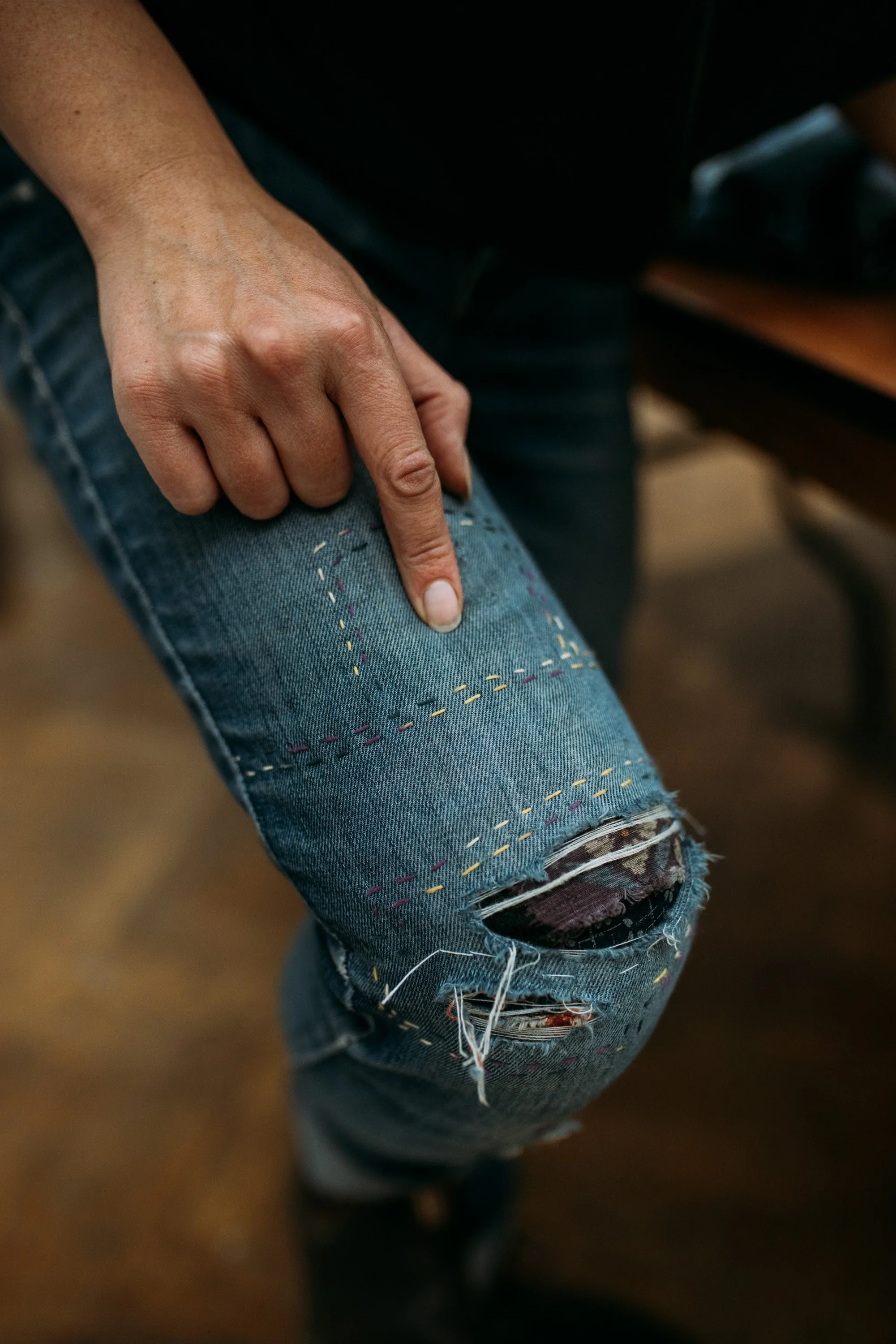 Person's hand pointing to a tear in their ripped blue jeans, with multicolored stitching visible.