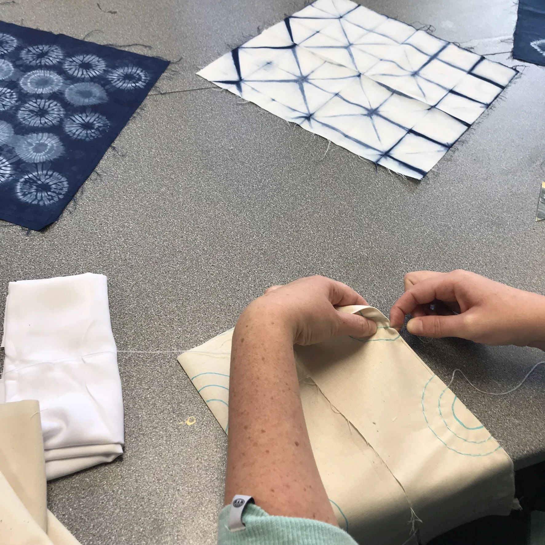 Person sewing white fabric with blue embroidered design, surrounded by patterned fabrics in blue and white, on a gray work surface.