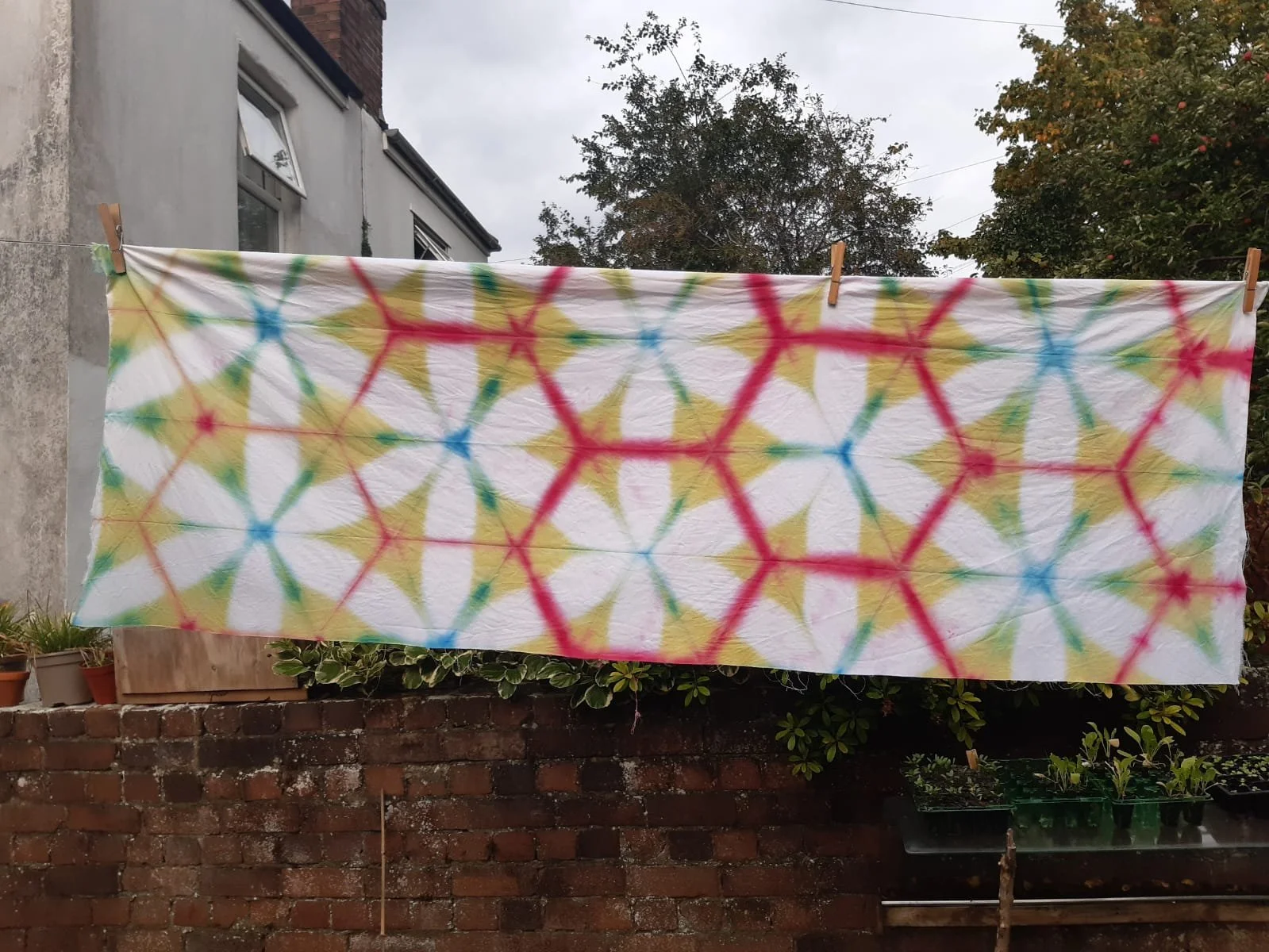 Colorful tie-dye cloth hanging on a line, secured with clothespins, outdoors against a sky and trees background.