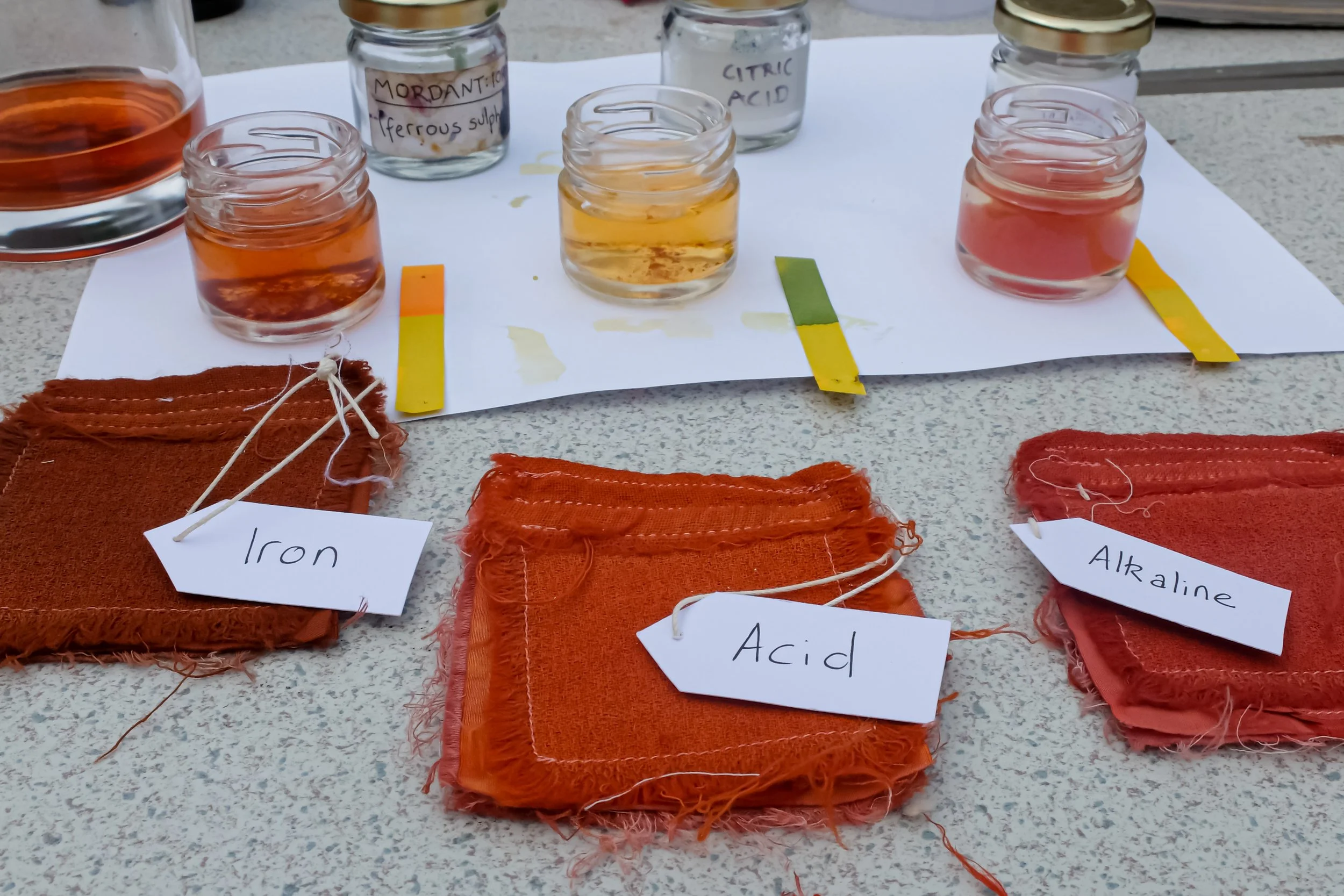 Samples of fabric labeled 'Iron,' 'Acid,' and 'Alkaline' arranged on a table with test solutions in small jars and color strips for chemical testing.