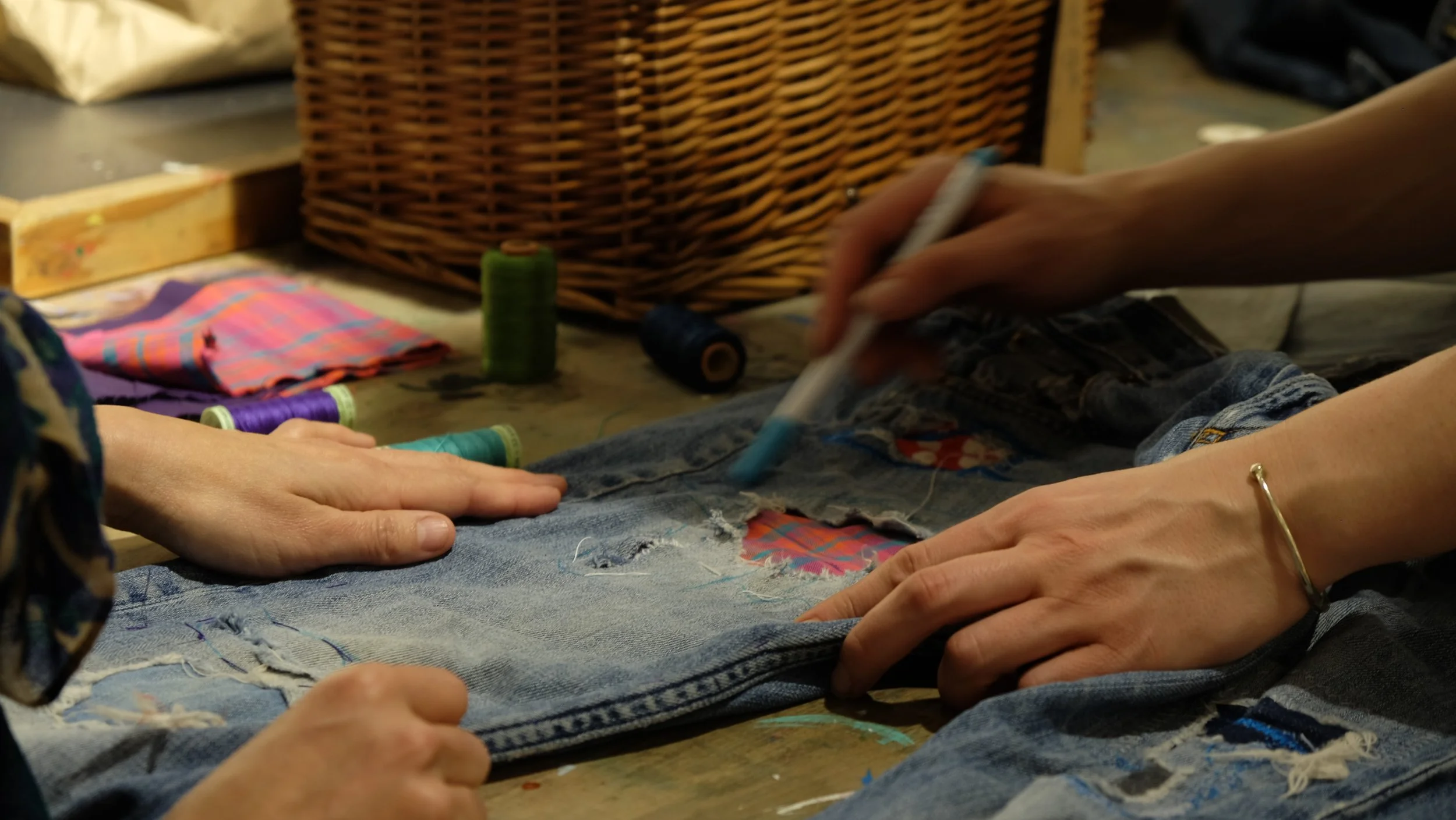 Hands sewing ripped denim jeans with colored threads on a worktable, surrounded by sewing supplies and spools of thread.