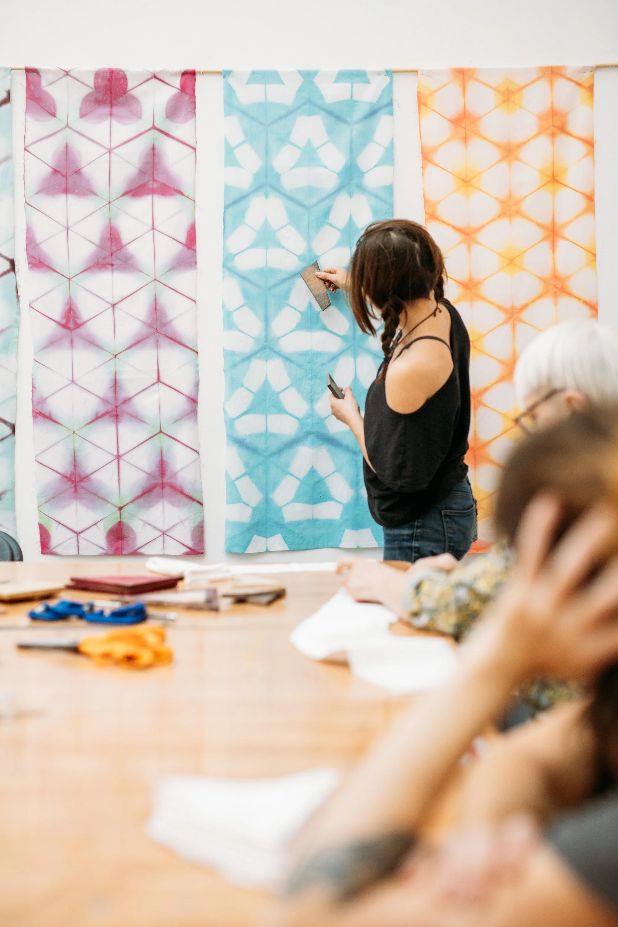 A woman standing in front of three colorful tie-dye fabrics hung on a wall, holding a tool while working on the fabric. Other people are seated at a table in the foreground, with craft supplies and papers on the table.