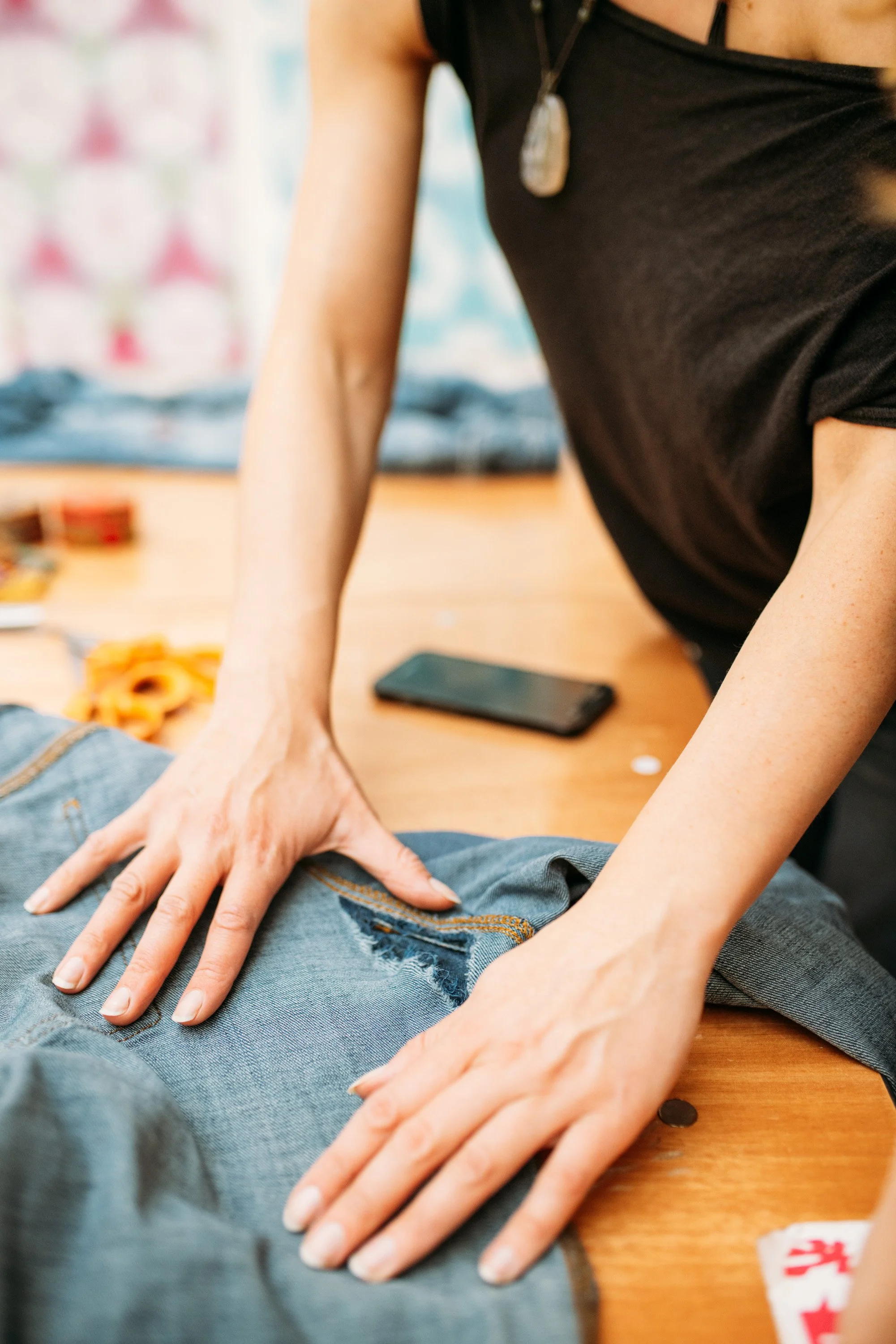 Person performing CPR on a pair of jeans laid flat on a wooden table, with a smartphone and snacks in the background.