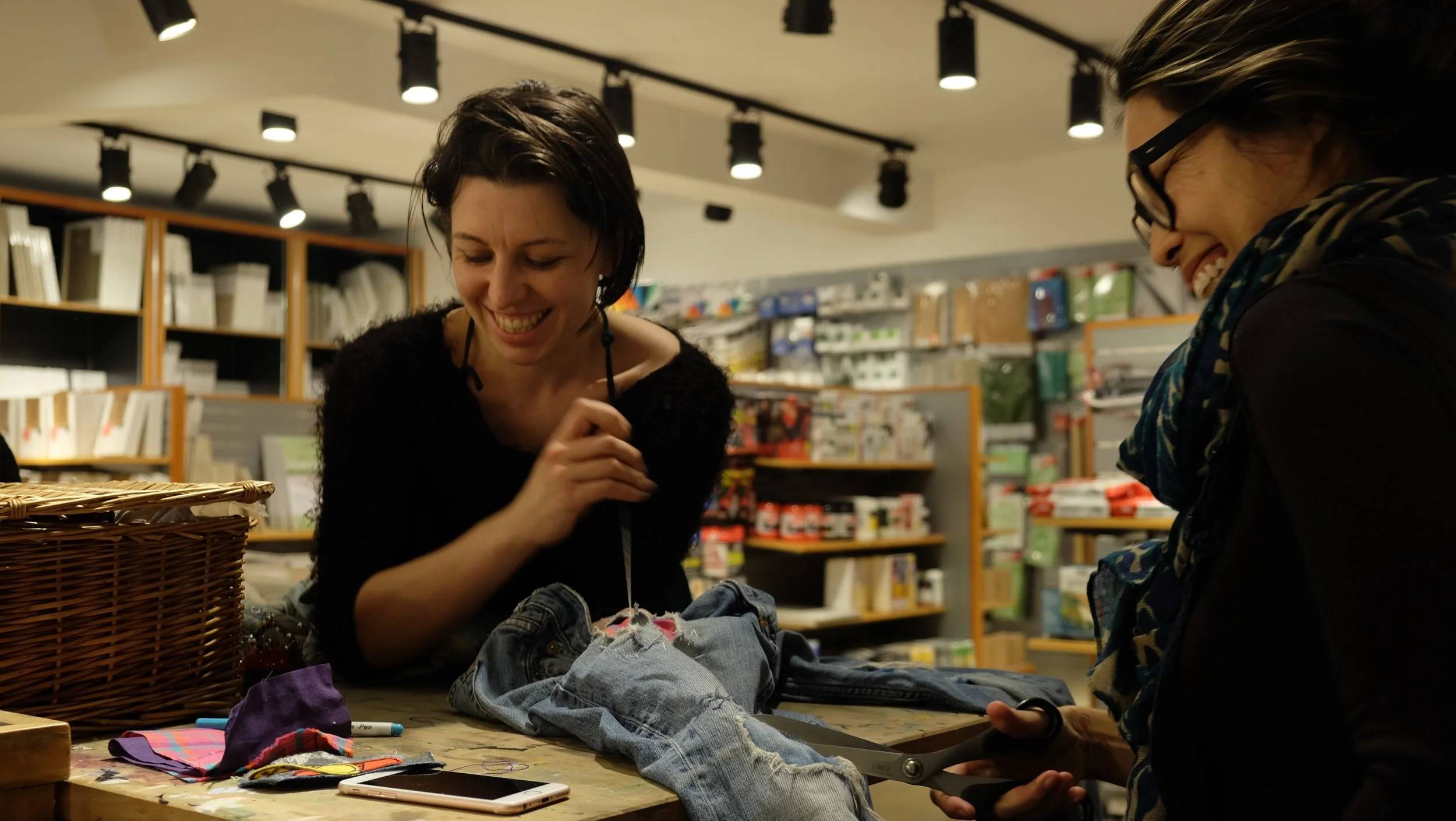 Two women smiling and laughing at a craft or sewing table in a store. One woman is holding a pair of scissors and working on a pair of jeans, while the other holds a smartphone. The table has fabric, markers, and a basket, with shelves filled with va