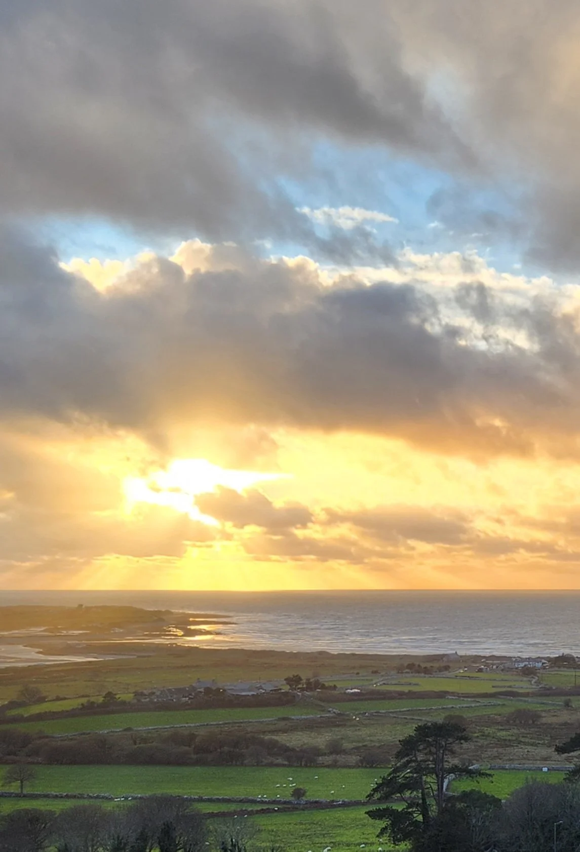 Scenic view of a sunset over the sea from Llanfair Slate Caverns, on the north west coast of Wales, UK with a partly cloudy sky and green fields in the foreground.