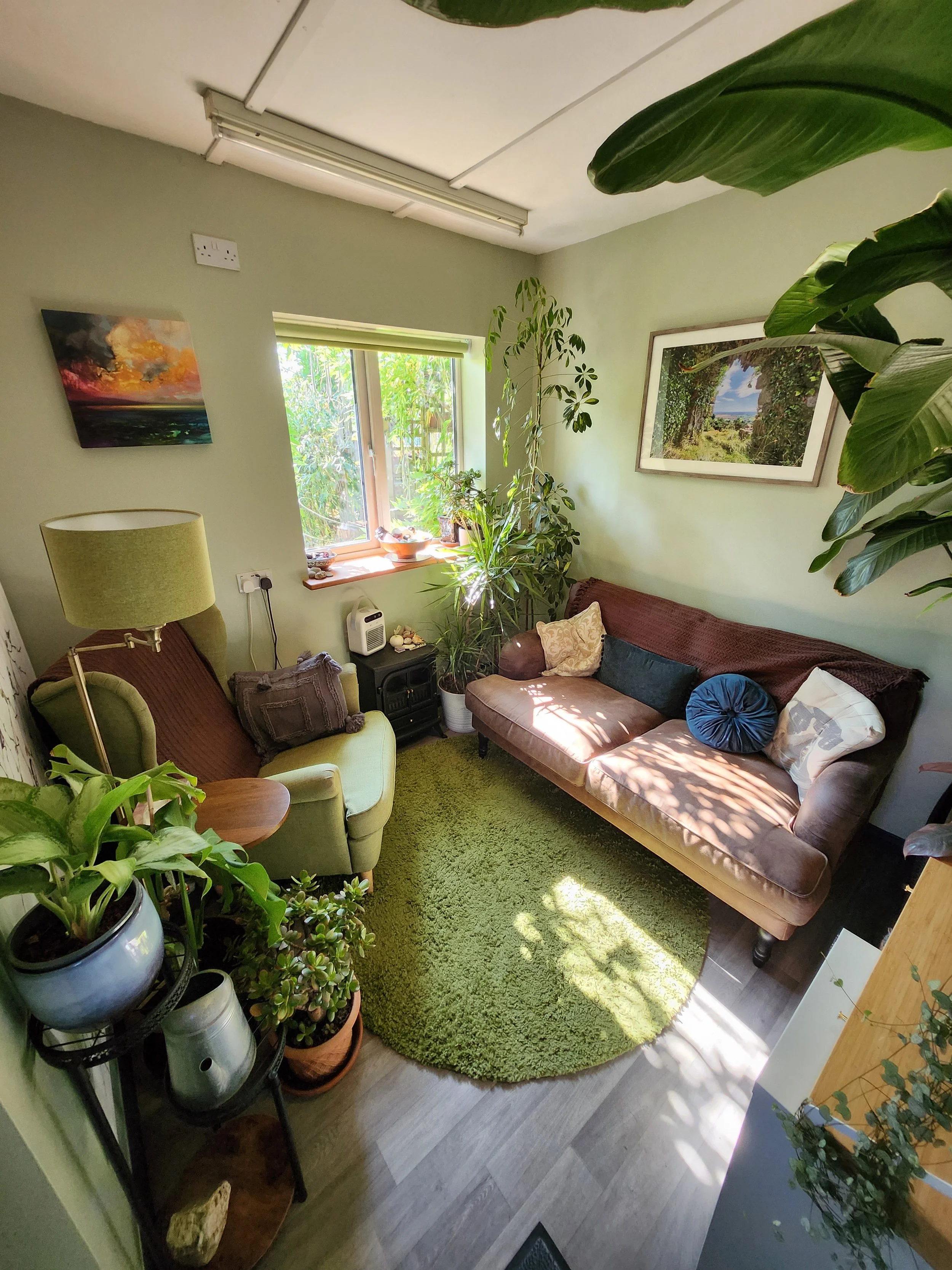 A cozy room with green walls and window allowing natural light. The room has a green armchair, a brown sof, and a green shaggy rug on the floor. Several potted plants decorate the space, including on a black metal plant stand. Artwork hangs on the walls, including a colorful abstract painting and a framed landscape photo.