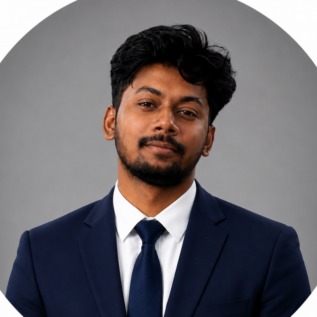 A young man with dark hair and a beard wearing a navy suit, white shirt, and navy tie, smiling against a white background.