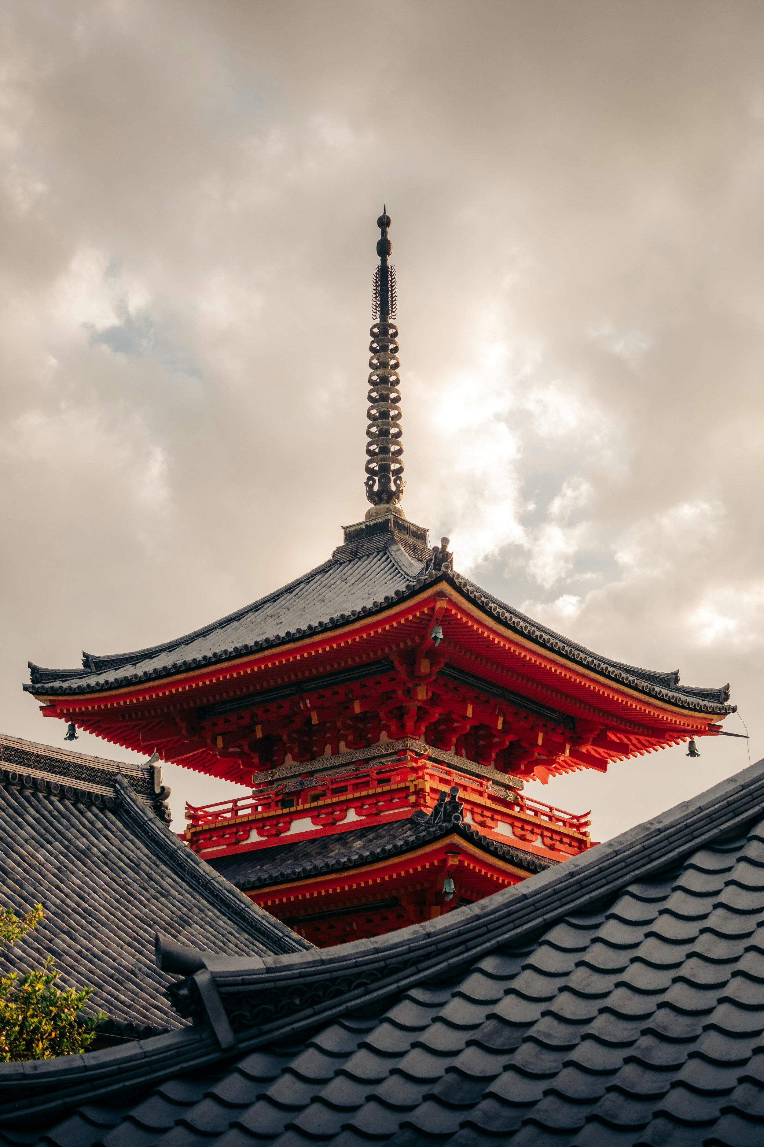 Traditional Japanese temple roof with a red pagoda structure and a tall, ornate spire extending into cloudy sky.