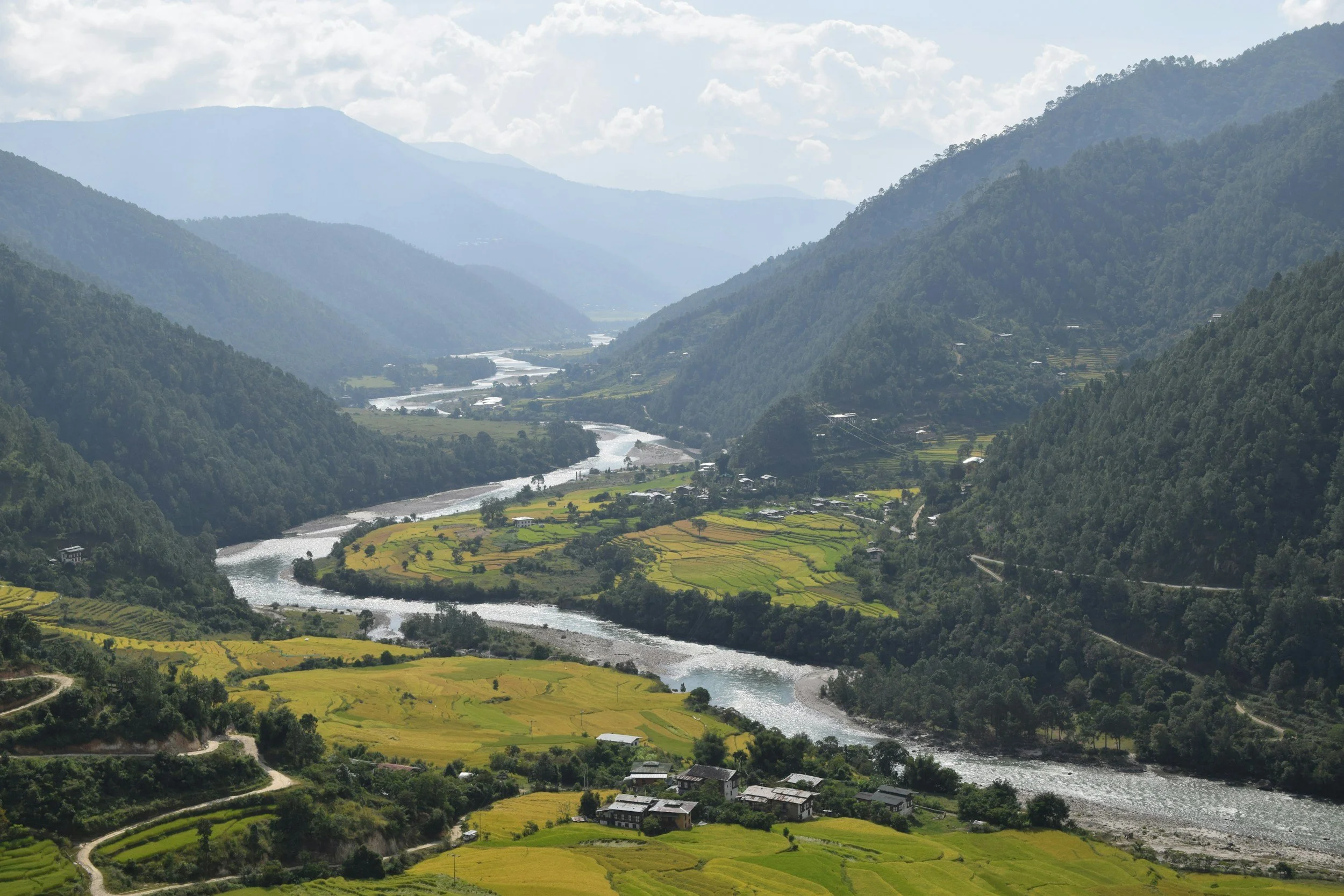 Scenic view of a river flowing through a lush green valley surrounded by tall mountains with dense forests, under a partly cloudy sky.