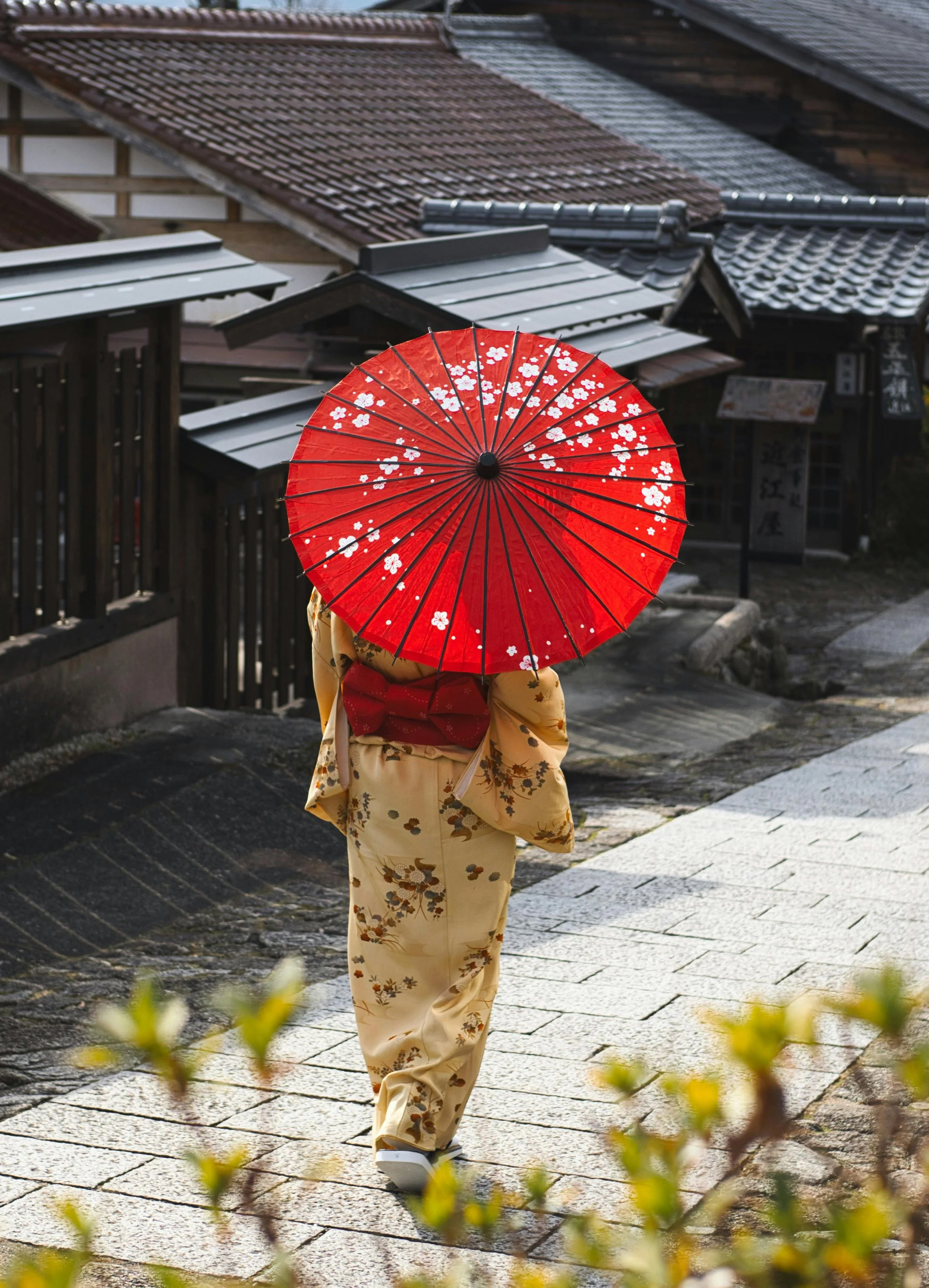 A person wearing traditional Japanese attire, holding a red parasol decorated with white cherry blossoms, walking on a stone street in a Japanese neighborhood with houses featuring tiled roofs.