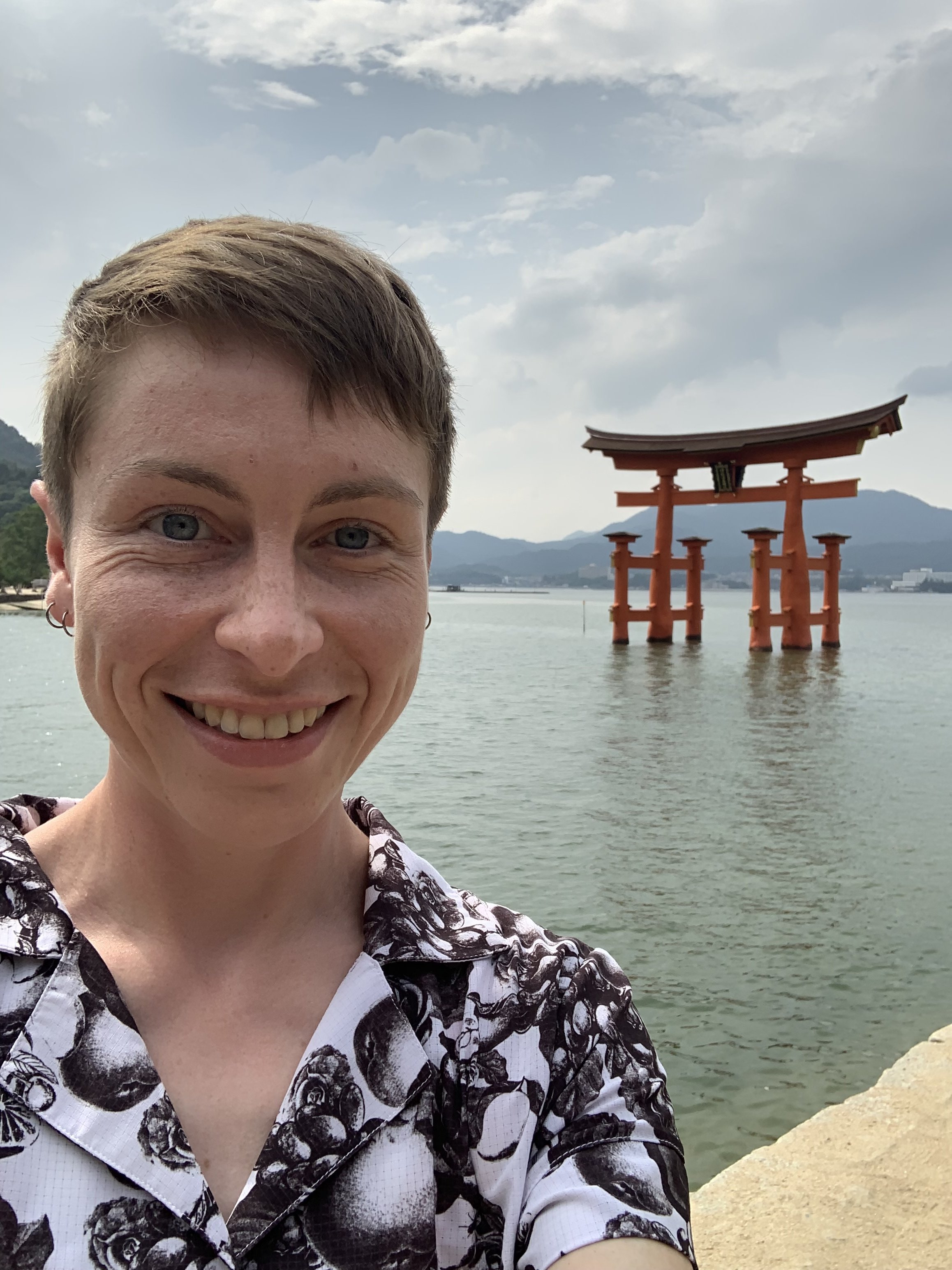 A woman with short brown hair and earrings smiling in front of a body of water. In the background is a traditional Japanese torii gate built over the water, with mountains and a cloudy sky behind.