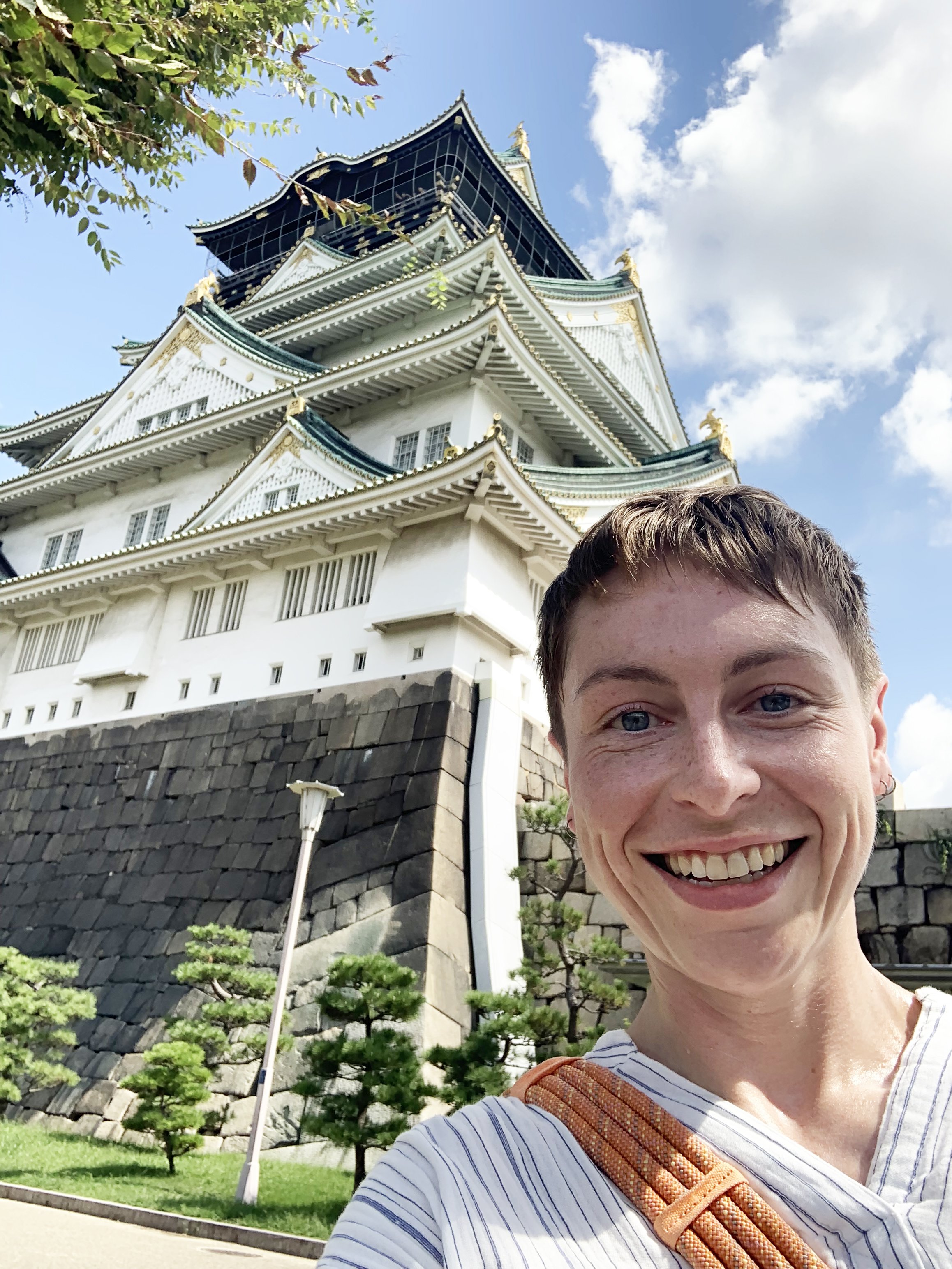 A smiling woman with short hair taking a selfie in front of a traditional Japanese castle with white walls and green roofs. The sky is partly cloudy and there are some trees and a lamppost visible in the background.