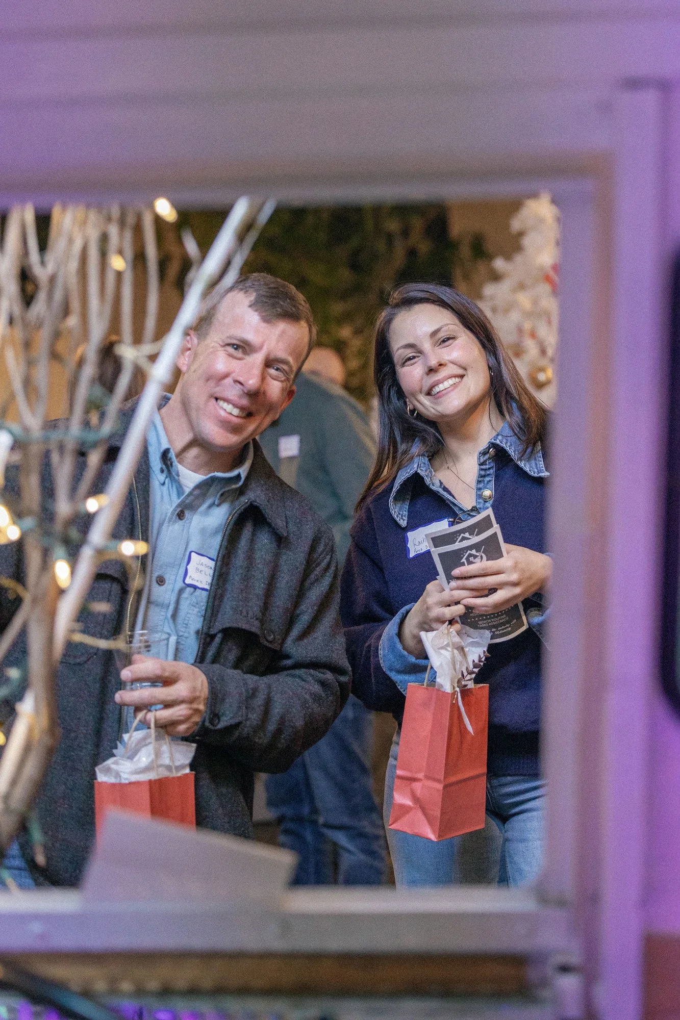 Two people smiling and holding gift bags at a holiday party, viewed through a window or frame.
