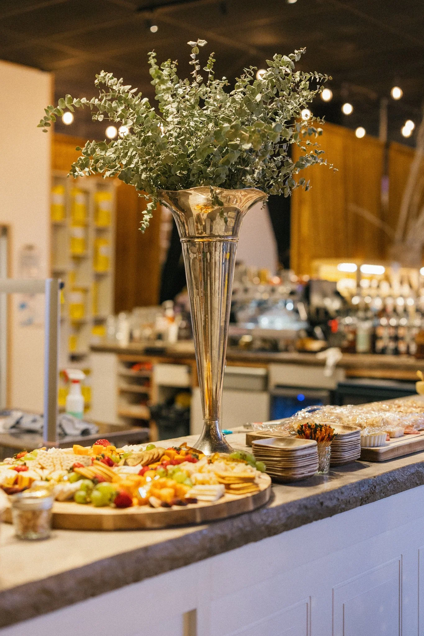 Decorative arrangement with a large jar filled with green eucalyptus leaves, surrounding a cheese and fruit platter on a bar or buffet table at a social event in a restaurant or cafe.