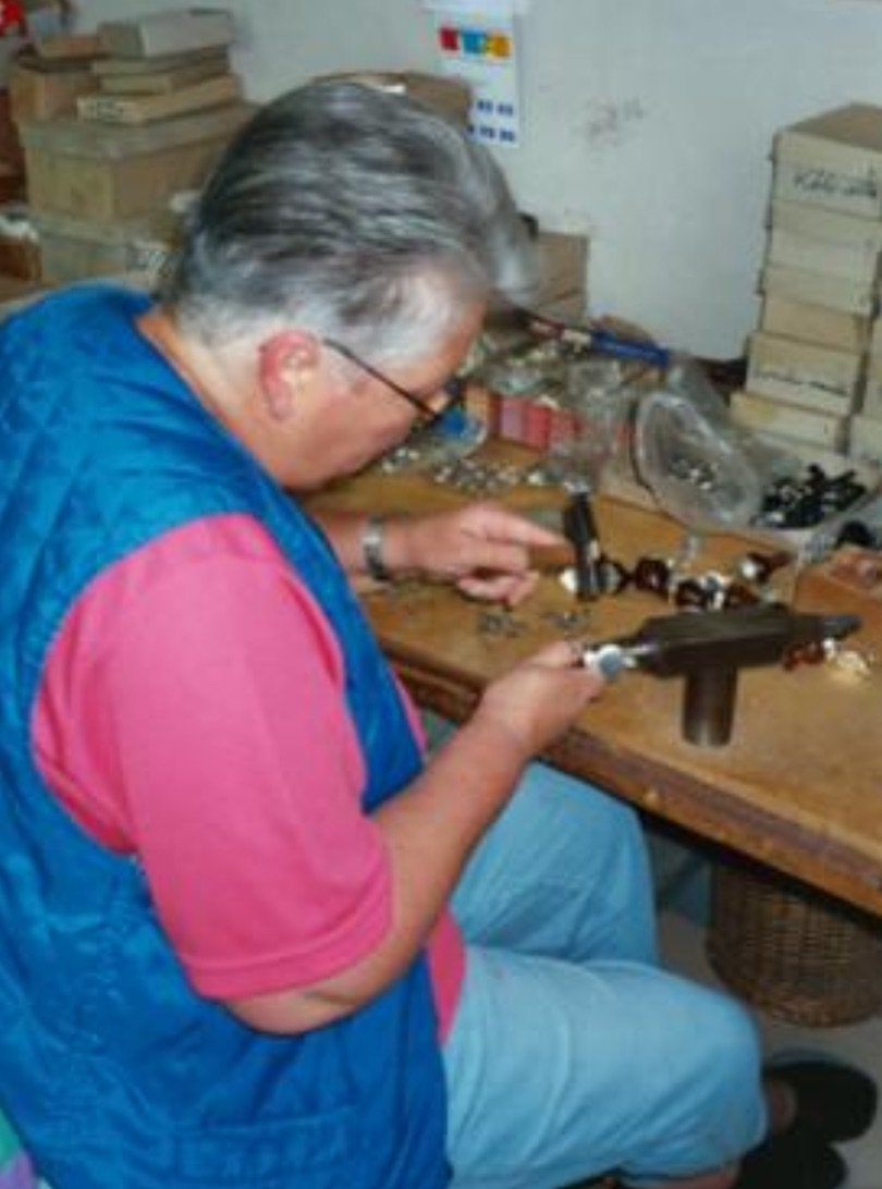 Ein Mann arbeitet an einem Basteltisch mit verschiedenen kleinen Gegenständen, vermutlich Werkzeuge oder Modellteile, in einem Raum mit Pappkartons im Hintergrund.