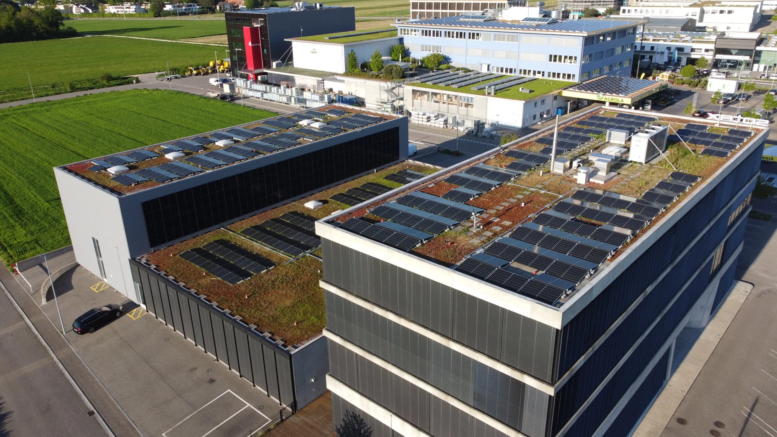 Luftaufnahme eines modernen Bürogebäudes mit grünem bepflanztem Flachdach, das mit Solarpanelen bedeckt ist, und umliegender grüner Landschaft sowie anderen Gebäuden.