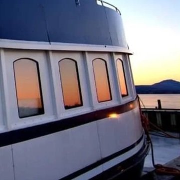 Close-up of a blue and white boat with windows reflecting a sunset over the water, docked at a pier.