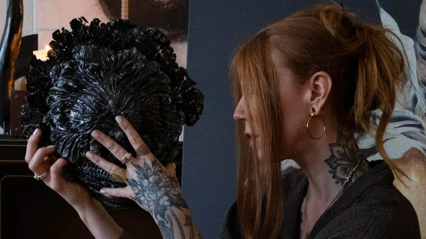 Eine Frau mit roten Haaren und Tattoos hält eine große schwarze künstlerisch gestaltete Skulptur vor ihr Gesicht.