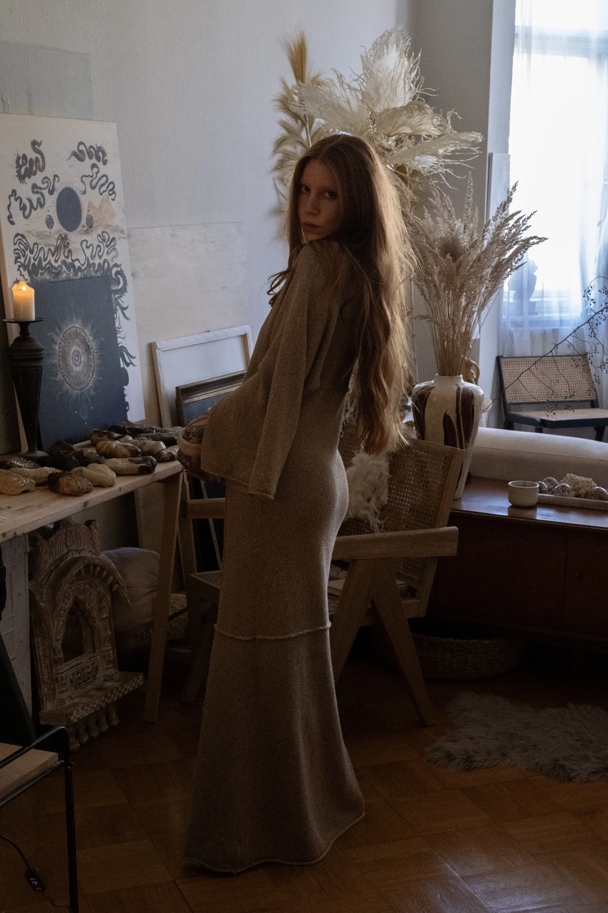Eine Frau mit langen roten Haaren steht in einem Atelier, das mit großen Trockenblumen und Kunstwerken dekoriert ist. Sie trägt ein beiges bodenlanges Kleid und schaut zur Kamera.