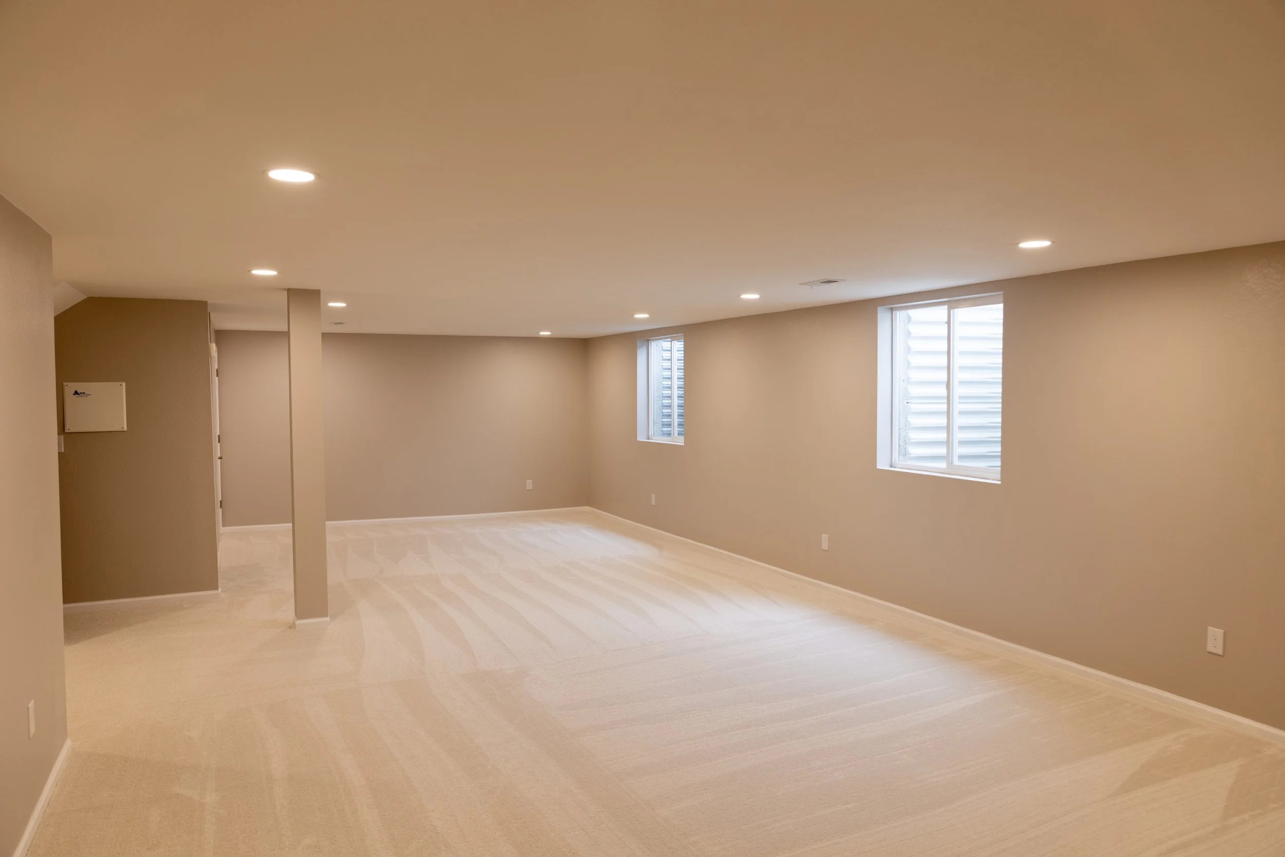 Empty basement room with beige walls, white carpet, recessed ceiling lights, and two small windows with blinds.