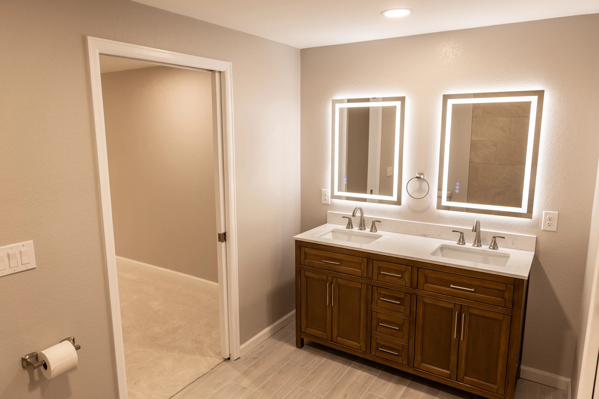 Bathroom with dual sinks, illuminated mirrors, wooden vanity, white countertop, beige walls, and a door leading to another room.