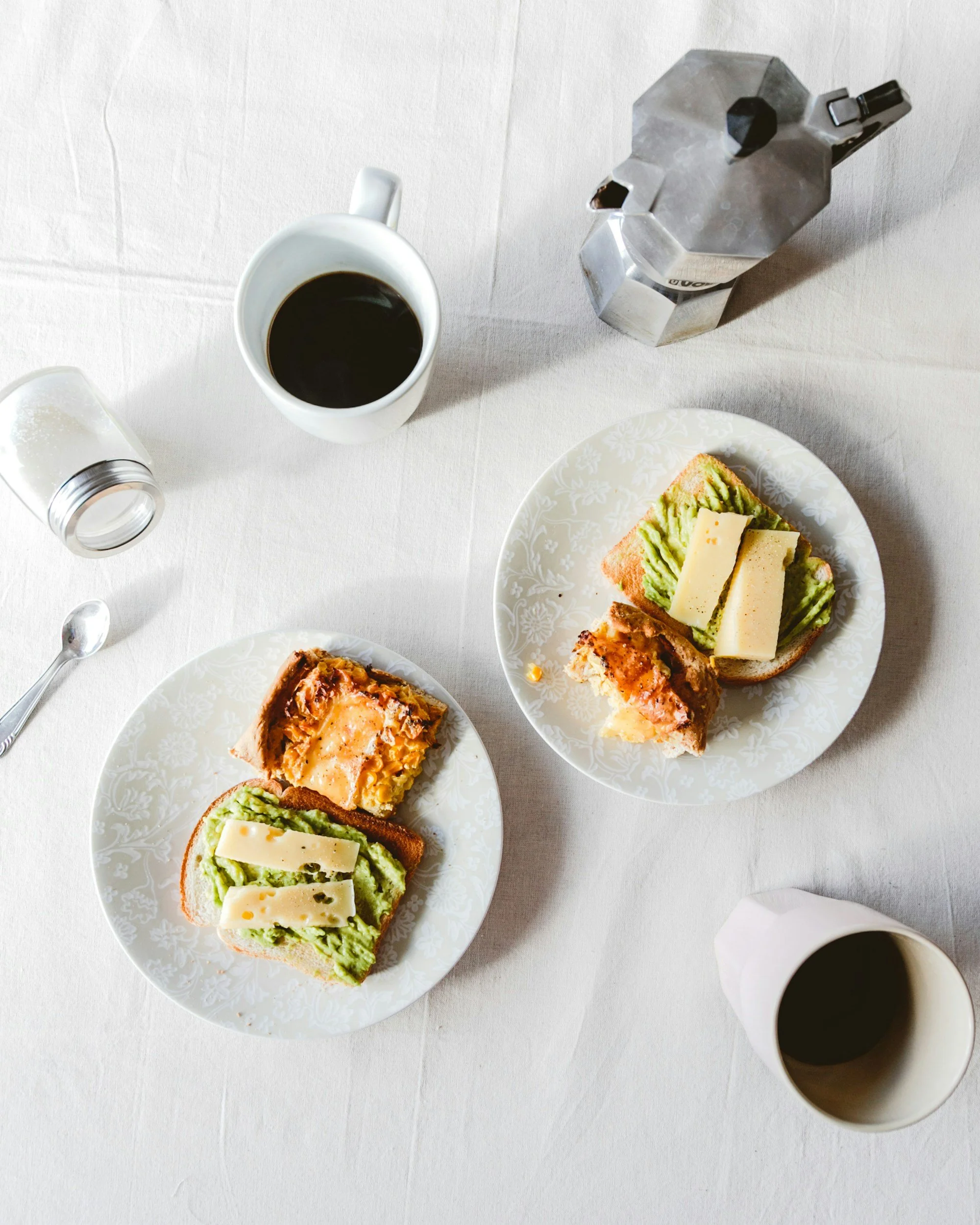 Top view of two plates with avocado toast with cheese, a slice of pizza, and green spread, a cup of black coffee, a glass of milk, and a moka pot on a white tablecloth