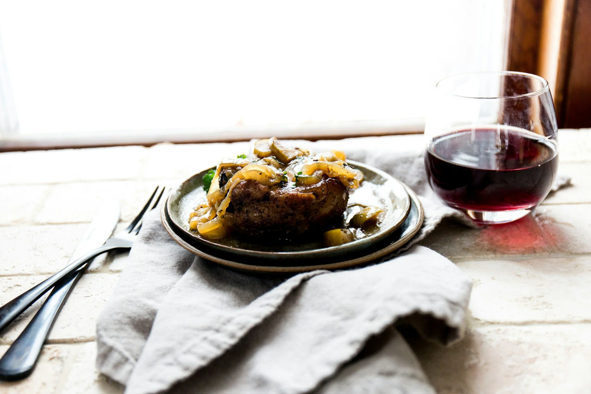 A plate with a cooked hamburger patty topped with sautéed mushrooms and onions, next to a glass of red wine on a white cloth napkin, with fork and knife on the side, set on a rustic wooden table near a window.