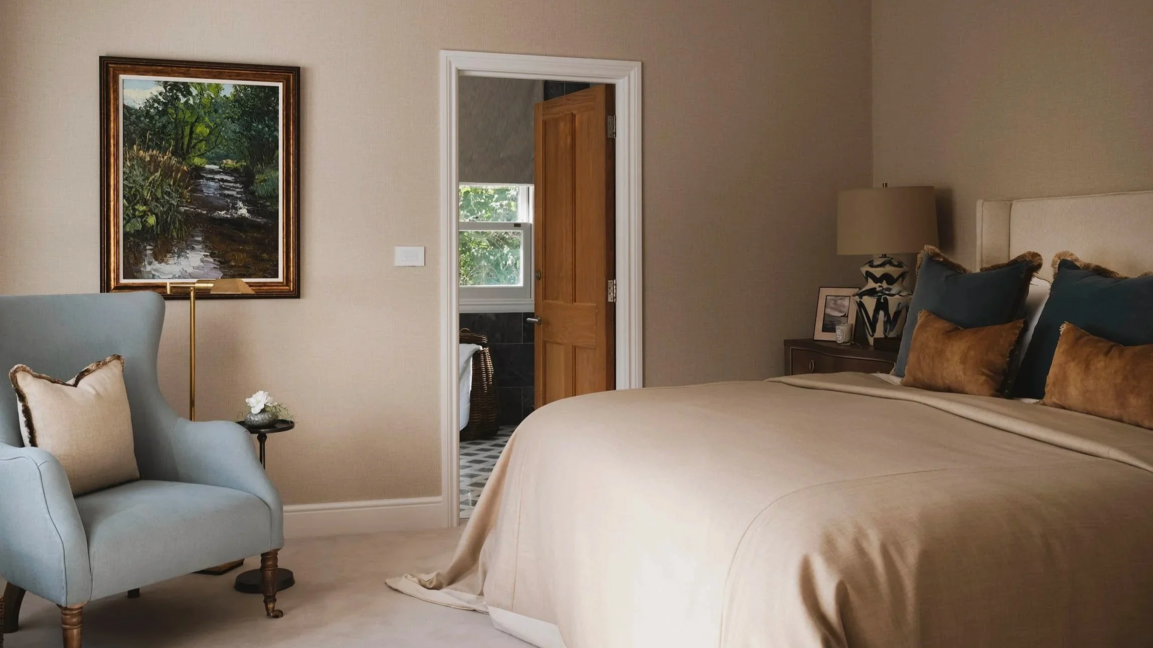 Luxury master bedroom suite with neutral linens, blue accent cushions, custom lighting, reading chair, and ensuite bathroom doorway.