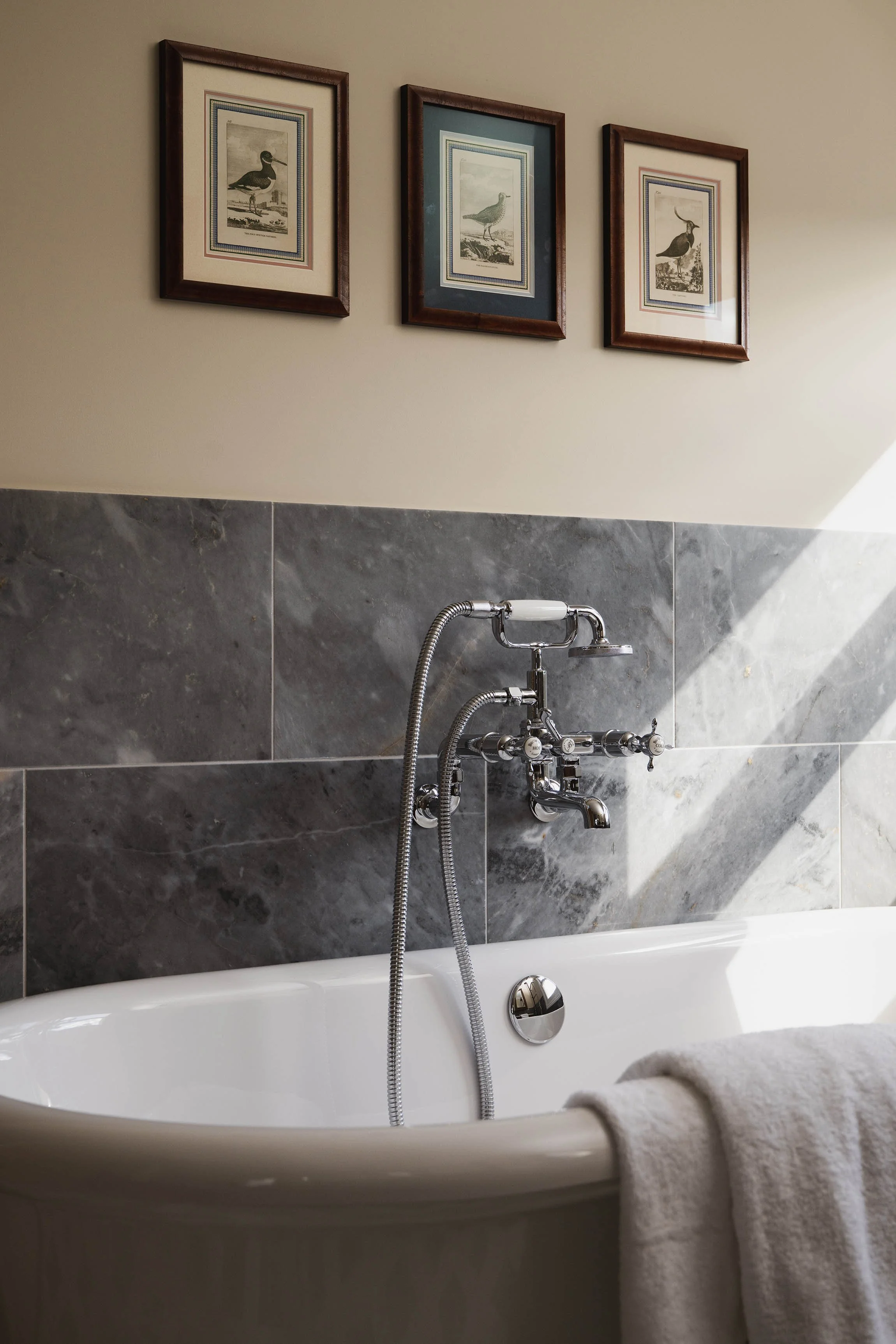 Freestanding soaking bathtub with traditional chrome taps, bluestone marble wall tiles, and framed bird prints.