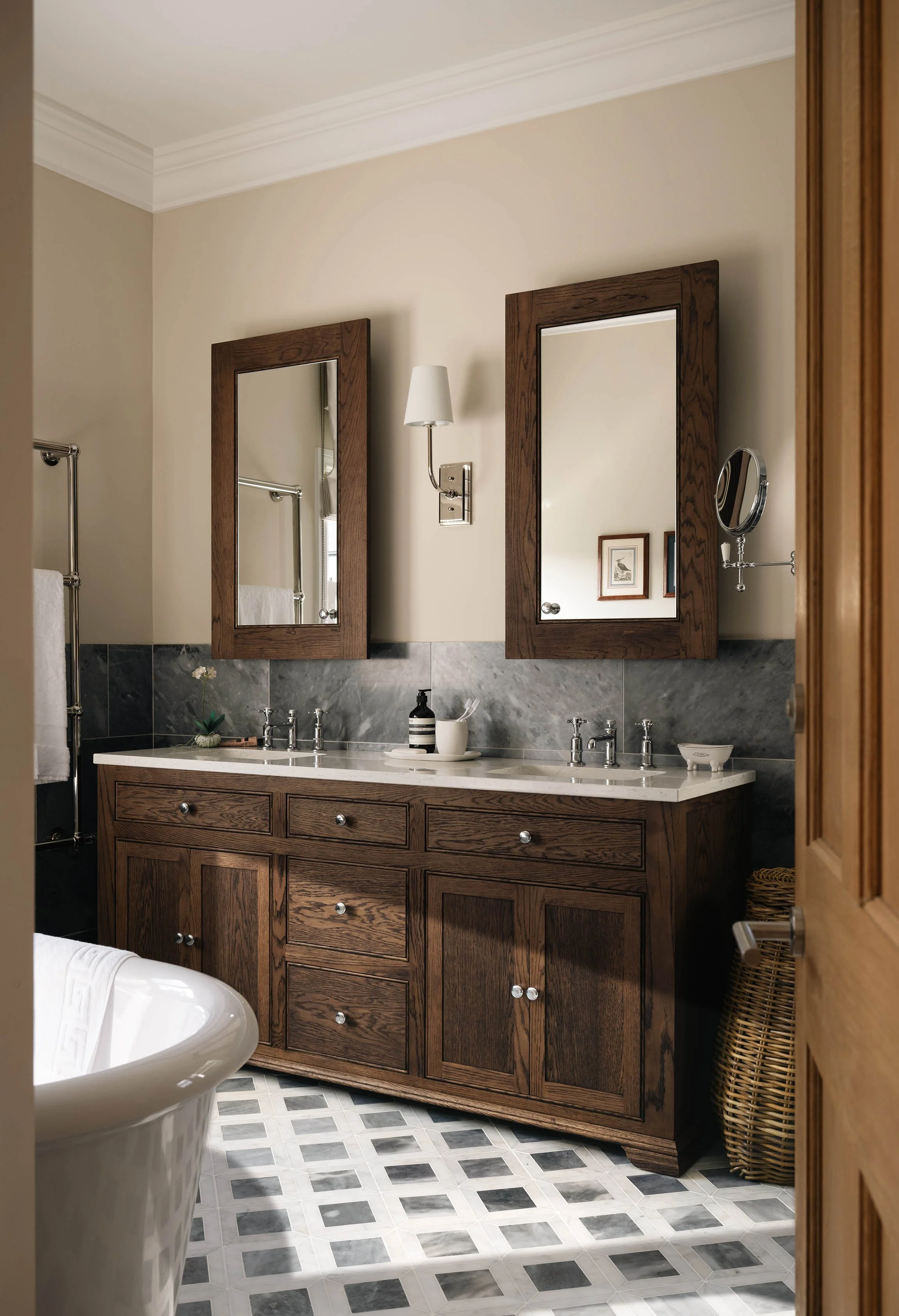 Traditional bathroom in a Woking, Surrey home, featuring a dark oak double vanity, framed mirrors, grey marble wall tiles and a classic checkerboard floor, designed by Joanne Quinn Interiors.
