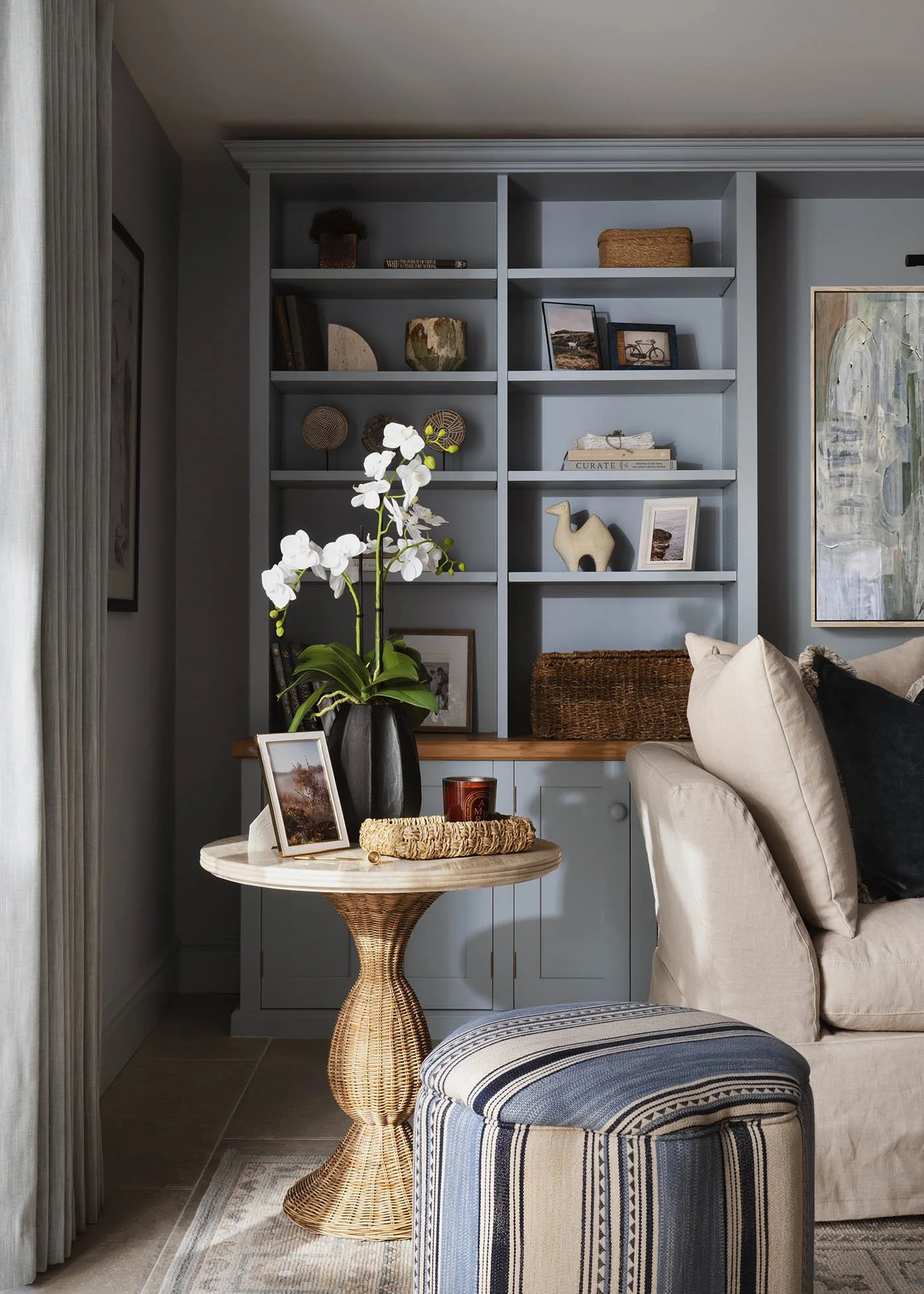 Elegant casual living room in a Woking, Surrey home, designed by Joanne Quinn Interiors, with a neutral, skirted sofa, pale blue walls, bespoke shelving, layered cushions and contemporary artwork.