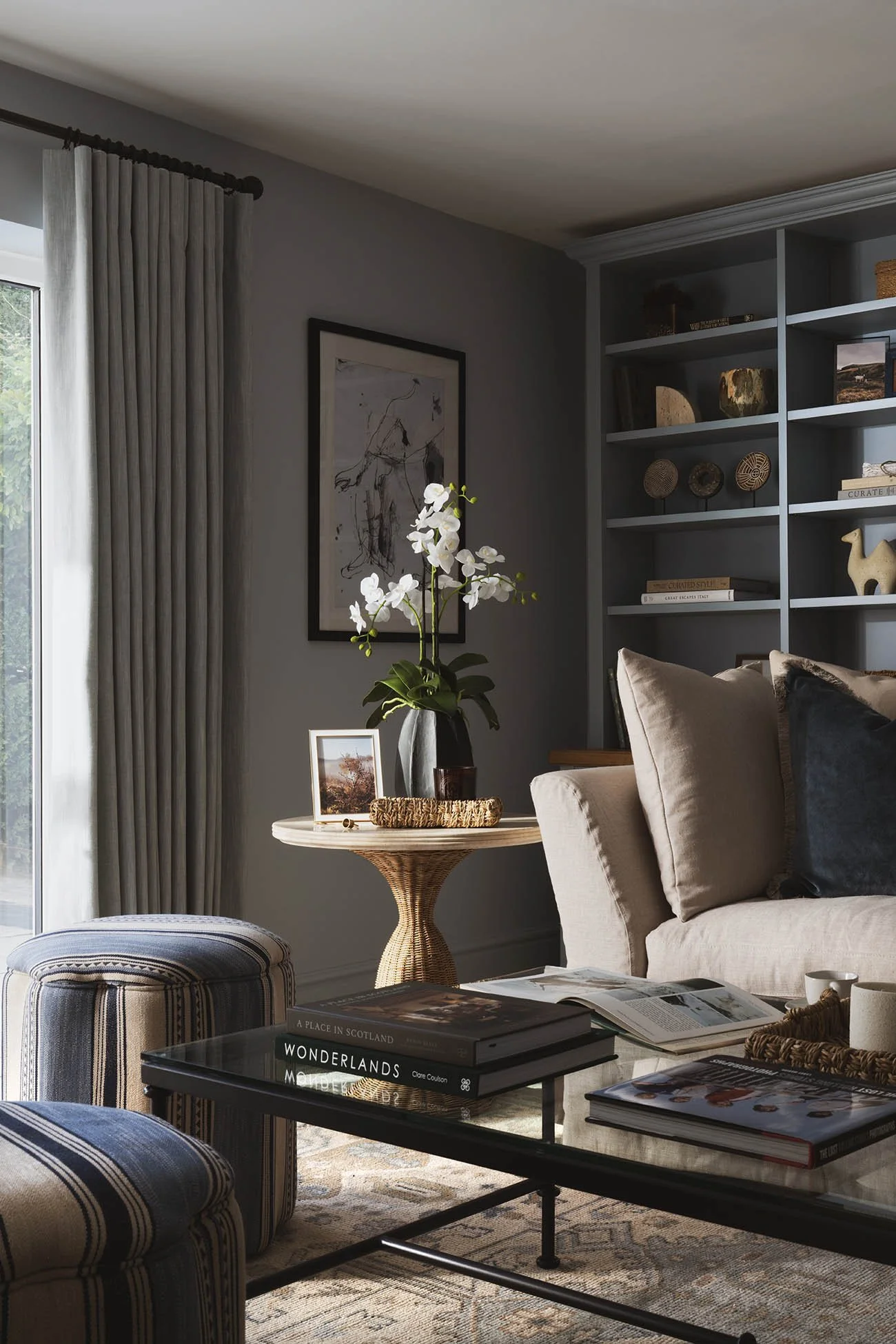 Elegant modern snug lounge with custom grey cabinetry, light neutral sectional sofa with velvet cushions, glass coffee table, and poolside views.