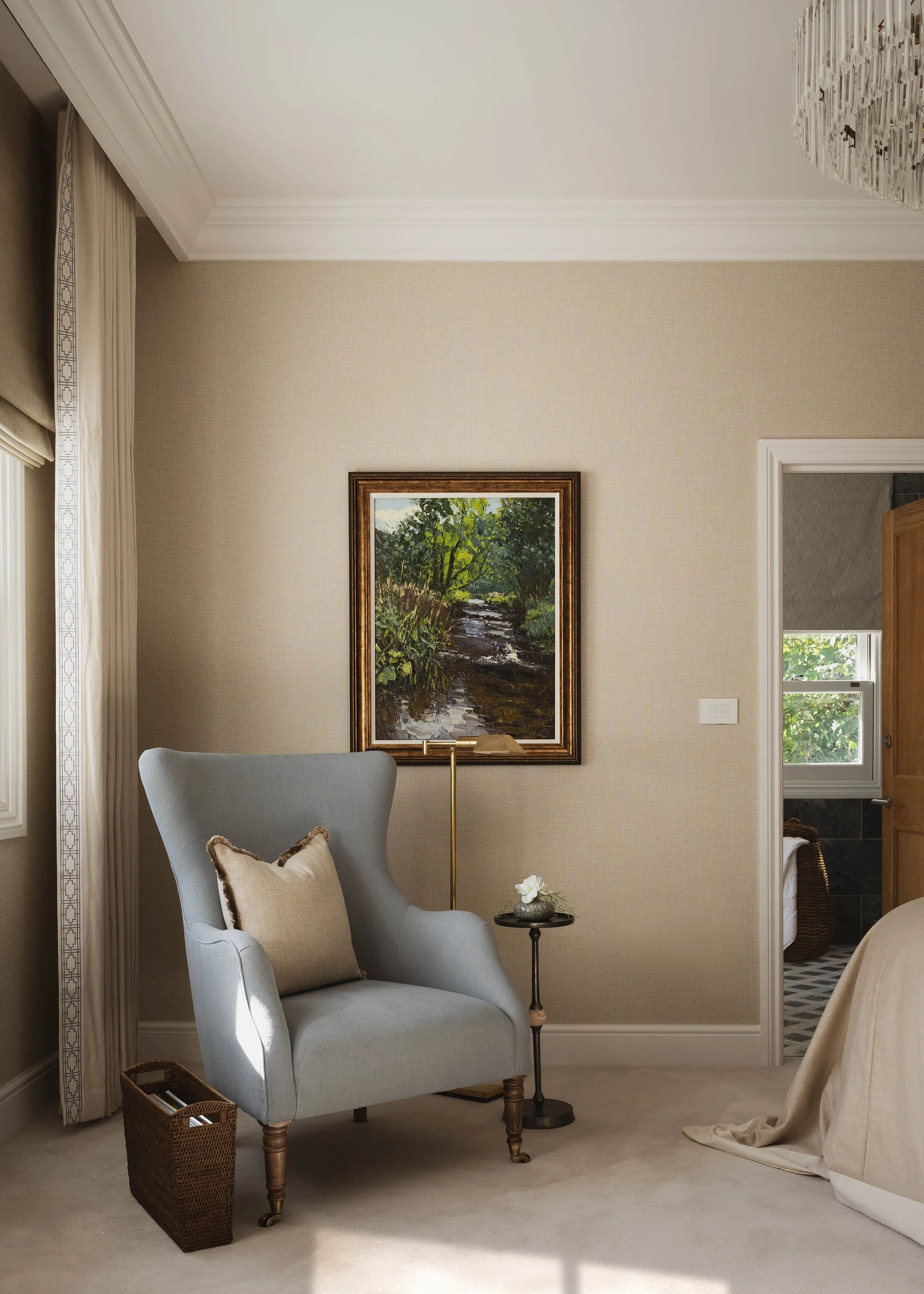 Master bedroom reading corner with a soft blue wingback arm chair, brass floor lamp, framed landscape art, and neutral textured wallpaper in a Woking residence.